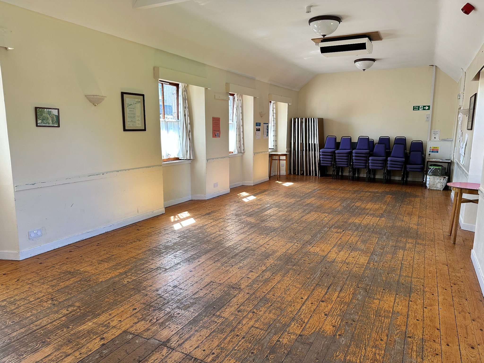 Empty room with wooden floor, cream-colored walls, three windows with curtains, and stacked purple chairs against the wall in the corner.