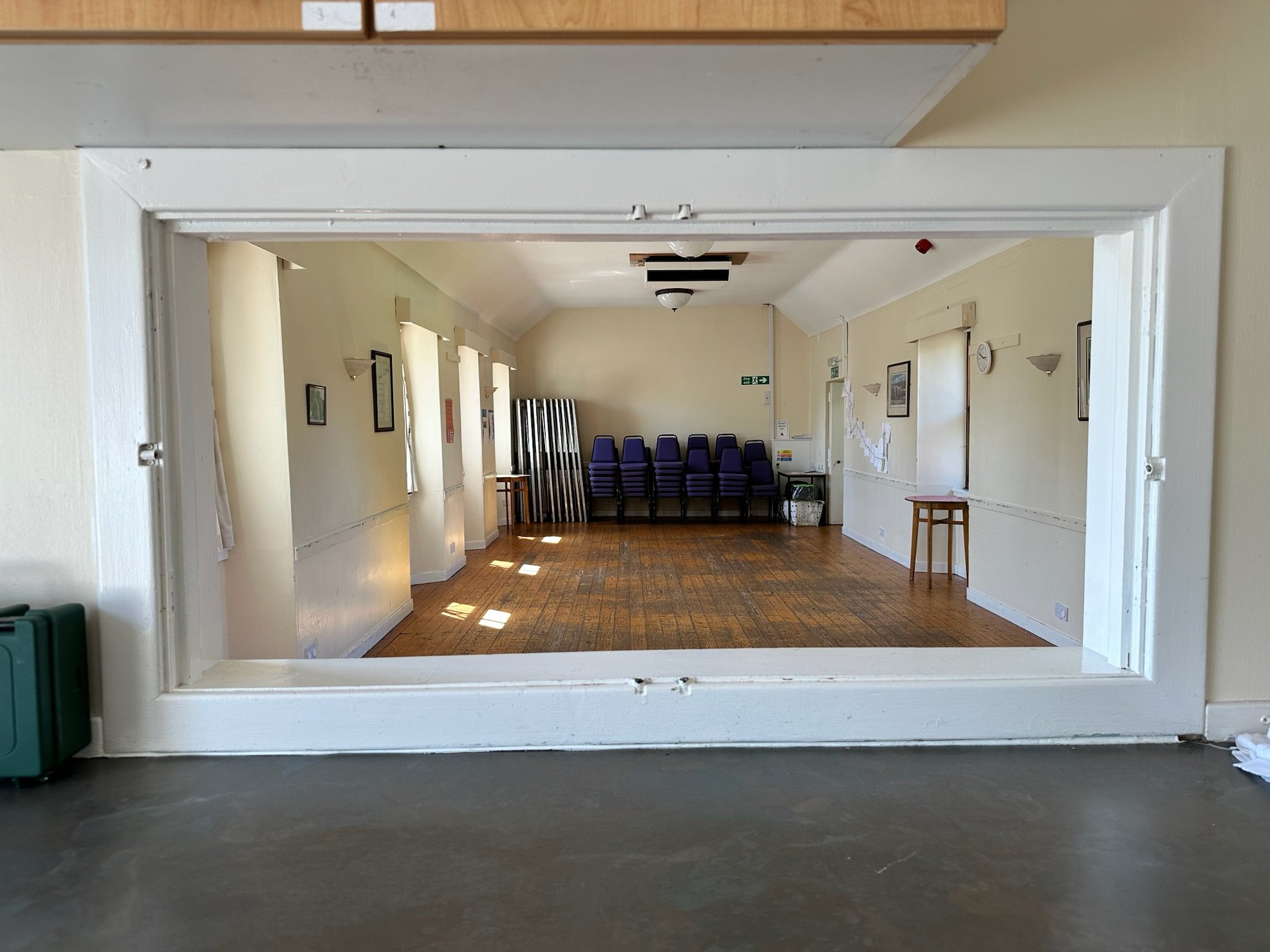 View of a room seen through a white window frame. The room has stack of purple chairs, a table, framed pictures on the walls, a clock, and some folded tables leaning against the back wall.