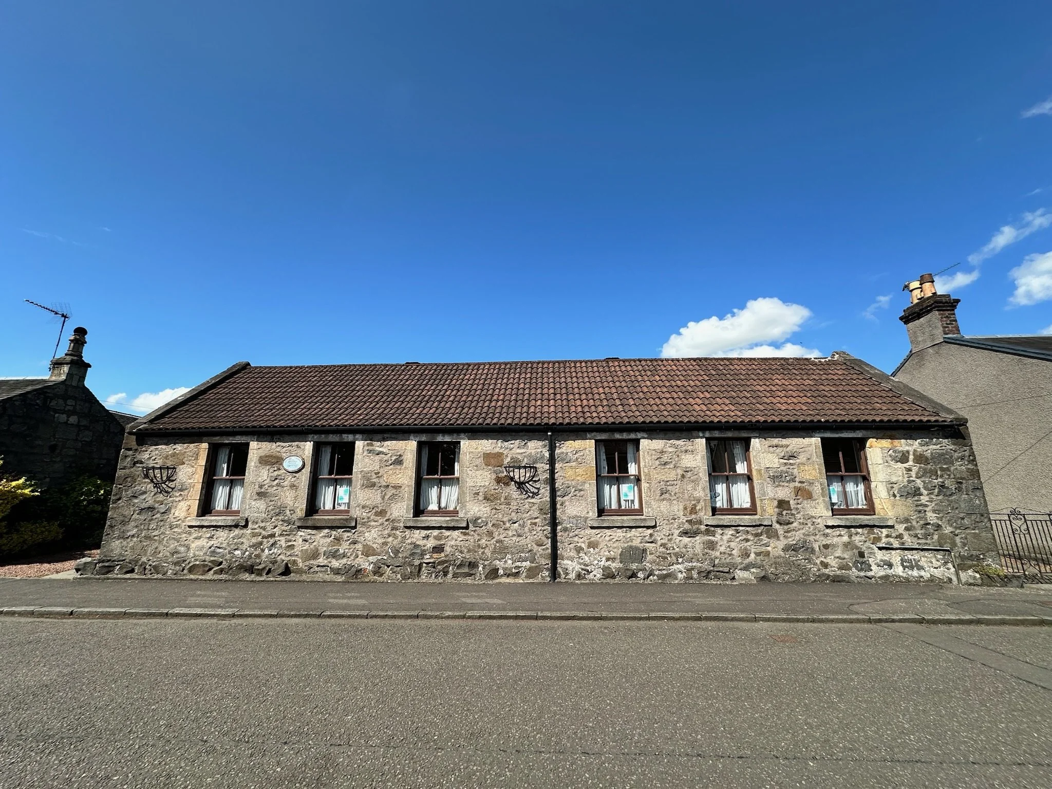 A stone building with a red-tiled roof, three windows with white curtains, and two chimneys against a blue sky with some white clouds.
