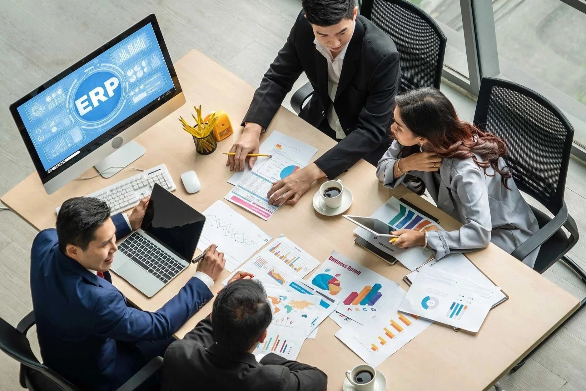 Business meeting taking place in a conference room with five professionals discussing graphs and data charts, surrounded by laptops, tablets, and printed charts.