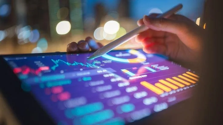 A person holding a stylus interacts with a digital tablet displaying financial charts and data, with a cityscape in the blurred background.