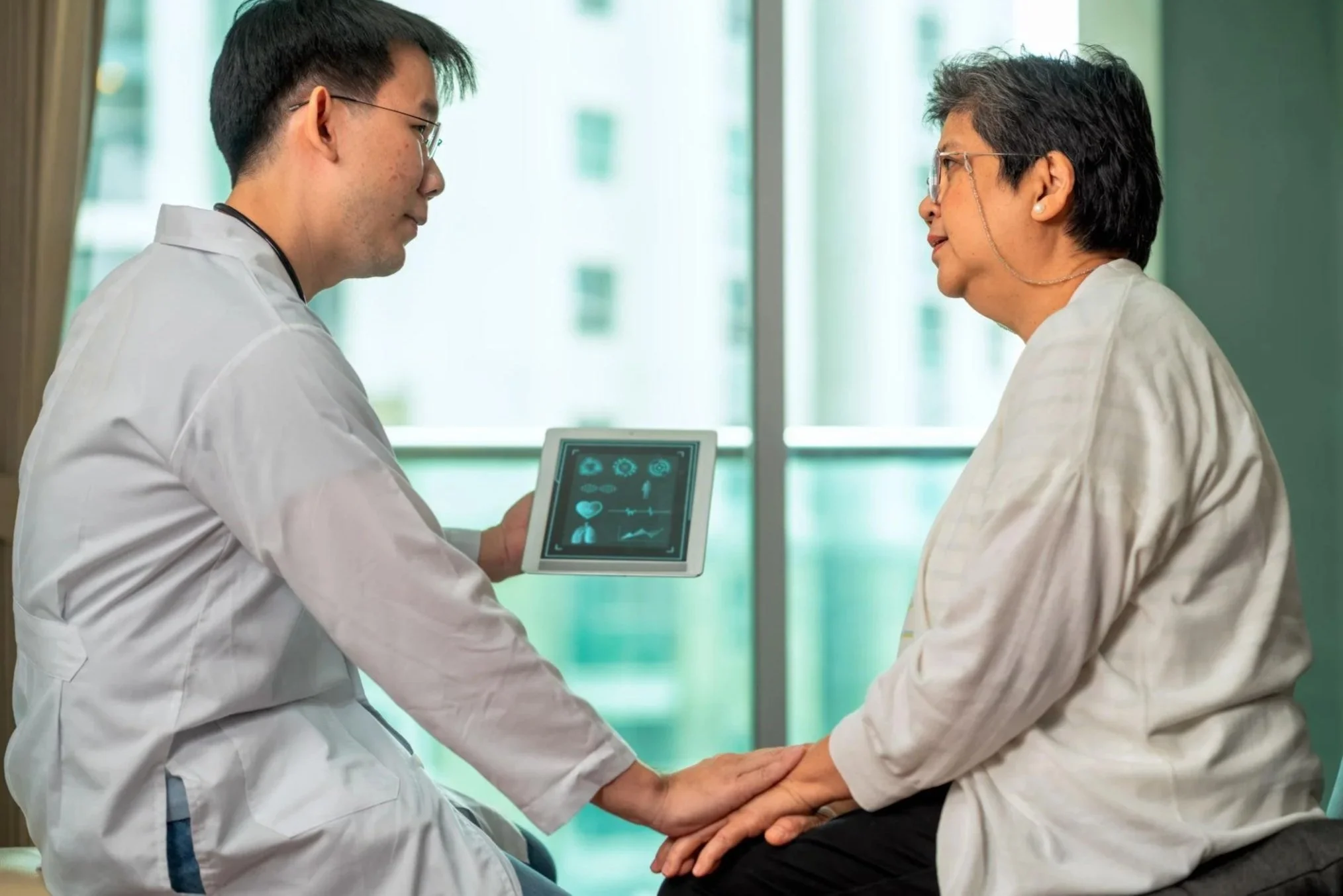 A male doctor and an elderly woman holding hands in a healthcare setting. The doctor is showing her medical information on a tablet, and they are engaging in a supportive conversation.