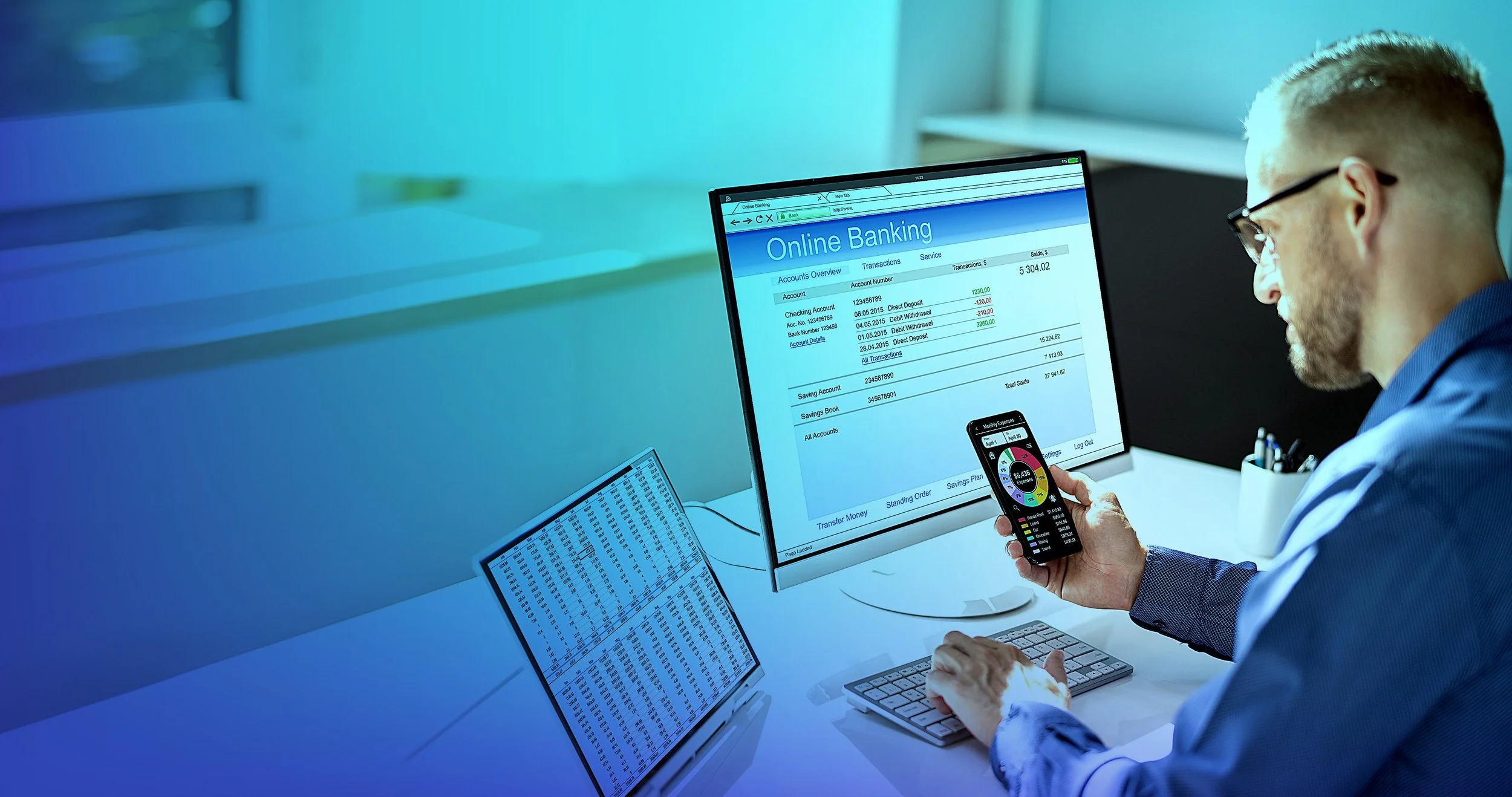 A man in glasses in a blue shirt using a smartphone with a colorful pie chart display while sitting at a desk with a computer monitor showing an online banking webpage. The webpage displays account overview, transactions, and balances. There is also a keyboard, a mouse, and a spreadsheet on the desk.