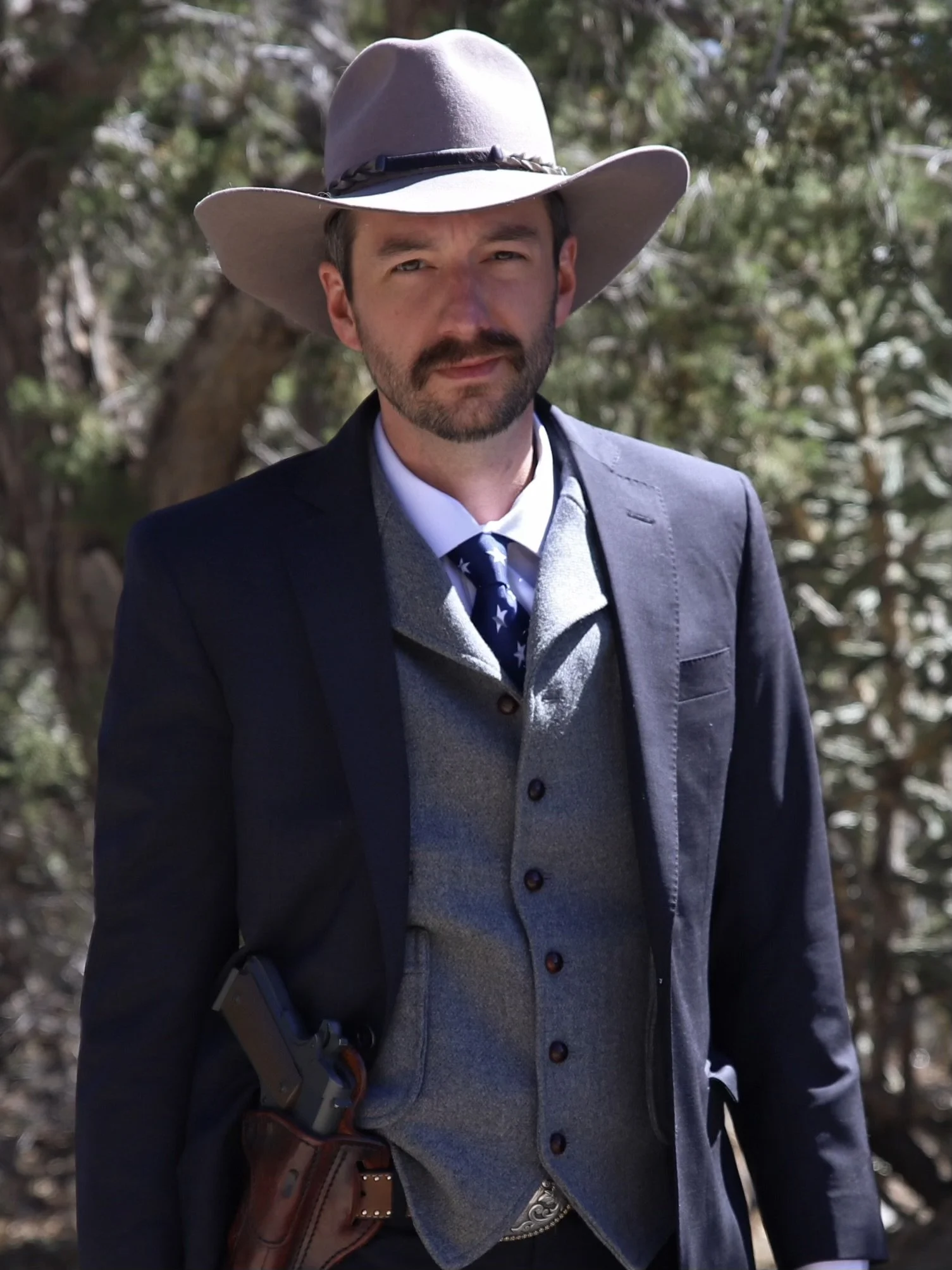 A man dressed as a cowboy standing outdoors in a wooded area, wearing a wide-brimmed hat, a suit jacket, a vest, and a tie, holding a revolver in a holster on his belt.