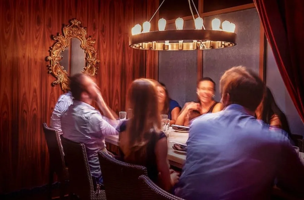 Group of friends sitting around a dining table in a cozy, dimly lit restaurant with wooden walls and red curtains, enjoying a conversation.