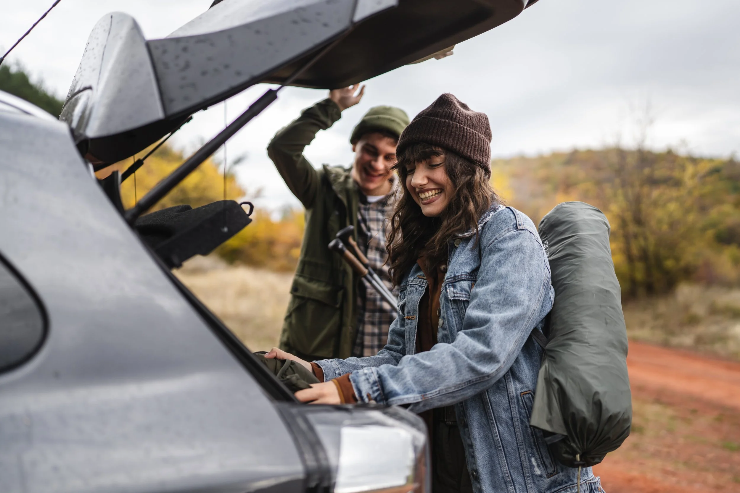 Two young adults, a man and a woman, are outdoors near a vehicle with its hood open, smiling and looking at the engine. They are dressed in casual autumn clothing, including beanies, jackets, and a backpack, with trees and fall foliage in the background.