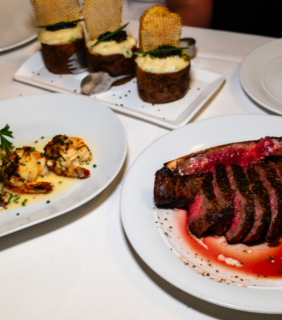A plate of sliced grilled steak with pink center on a white plate with juice, garnished with herbs; a plate with two seared shrimp and herbs in oil; and a tray of gourmet cannoli with cream filling, chocolate, and crispy wafers on a white tablecloth.