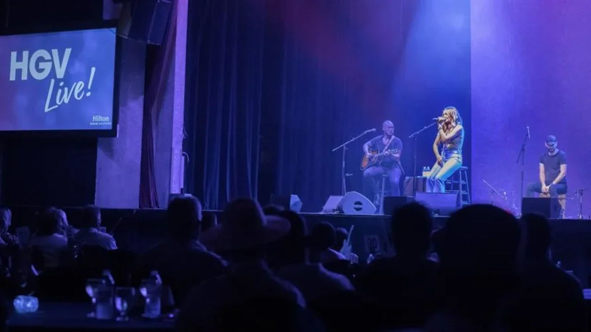 Live musical performance on stage with three performers, one playing guitar, one singing, and one sitting, in a dark theater setting, with an audience watching.
