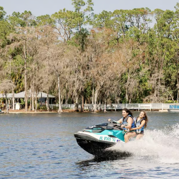 Two young women riding a jet ski on a lake with trees in the background.