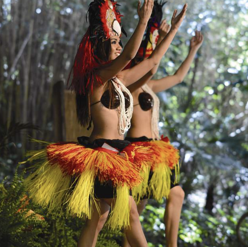 Two women wearing traditional tribal costumes with grass skirts, headdresses, and necklaces, dancing outdoors in a forest setting.