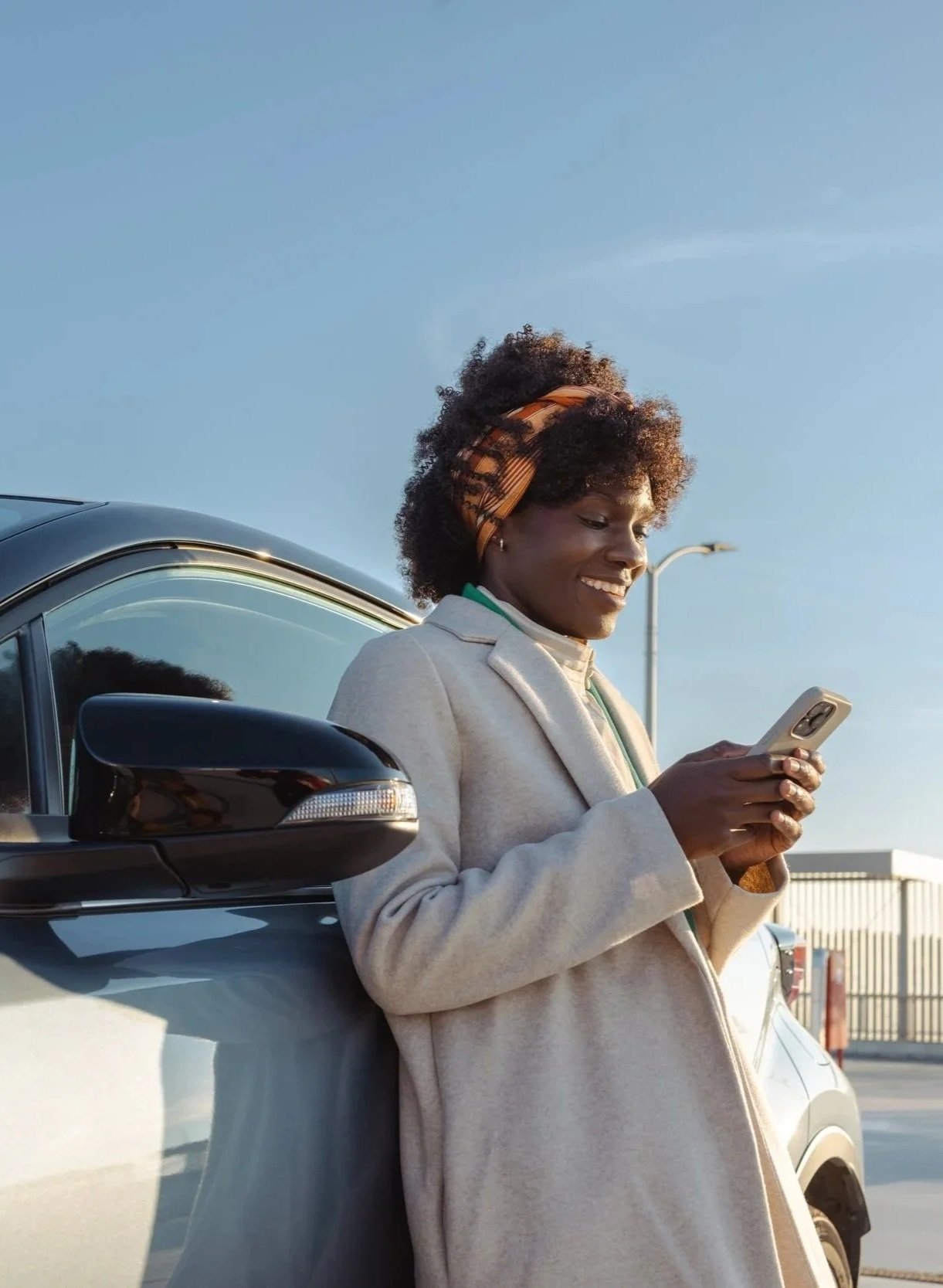 A woman with a headband standing outdoors next to a car, smiling and looking at her phone.