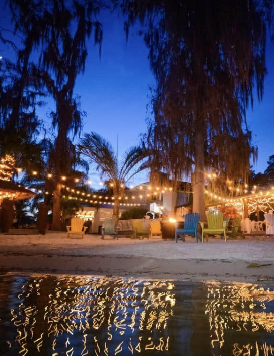 Outdoor beach scene at dusk with string lights, palm trees, colorful chairs, and a calm water reflection.