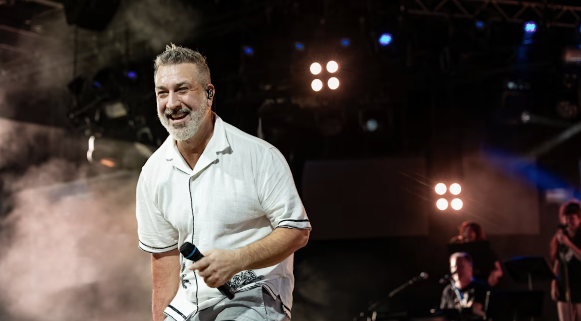 A man with gray hair and a beard, wearing a white shirt, performing on stage with a microphone in hand, smiling with stage lights and musicians in the background.
