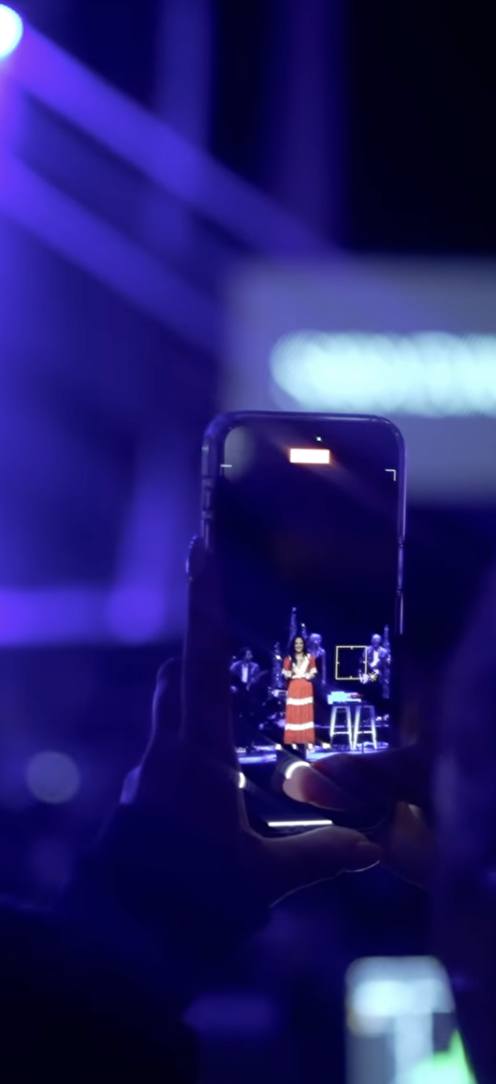 Person taking a photo of a singer on stage with a smartphone, stage lit with purple lights, singer in red dress.