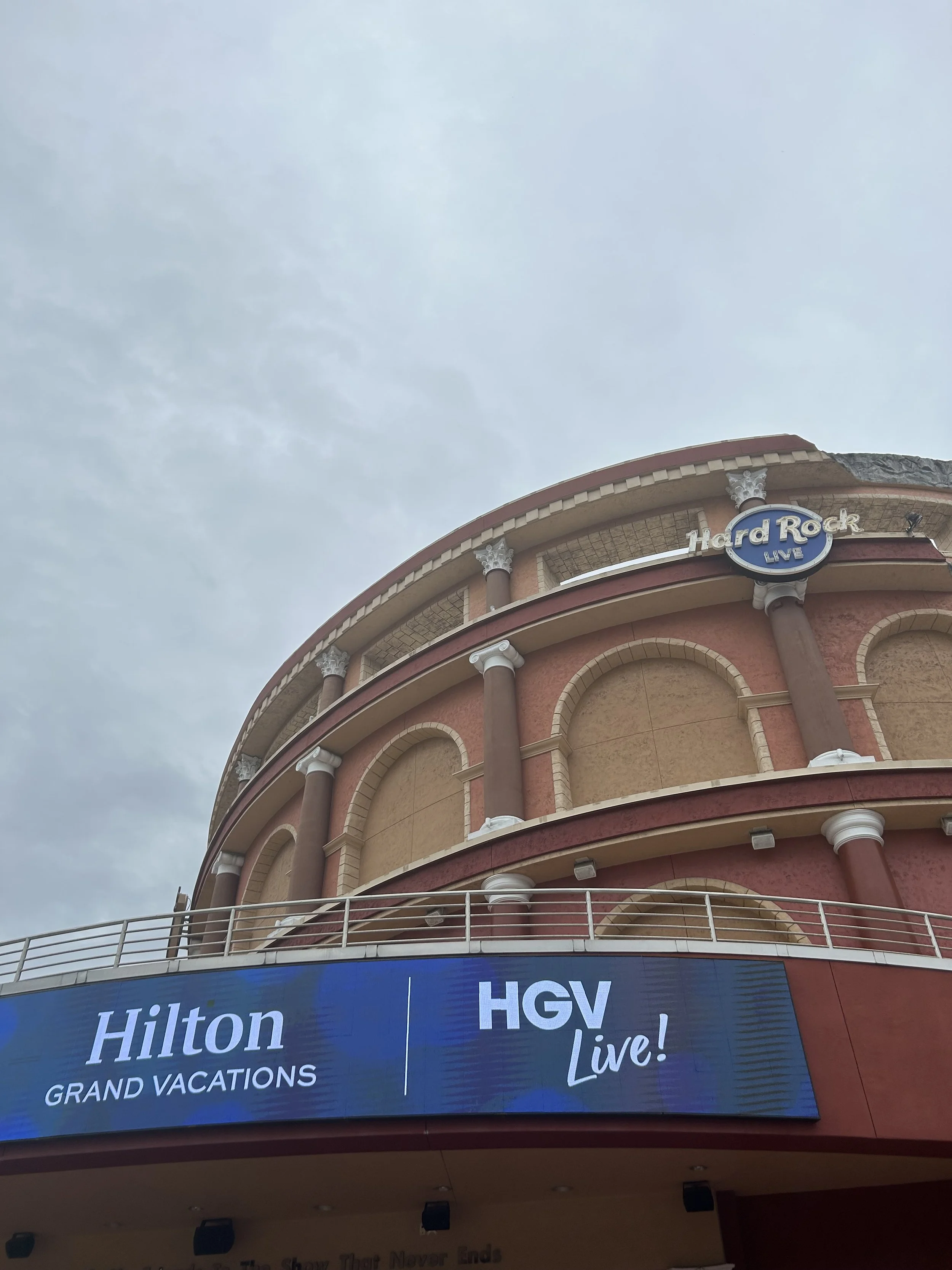 The photo shows the upper part of a multi-story building with a large sign that reads "Hard Rock Live". Below that, there's a digital billboard displaying the words "Hilton GRAND VACATIONS" and "HGV". The sky is partly cloudy.
