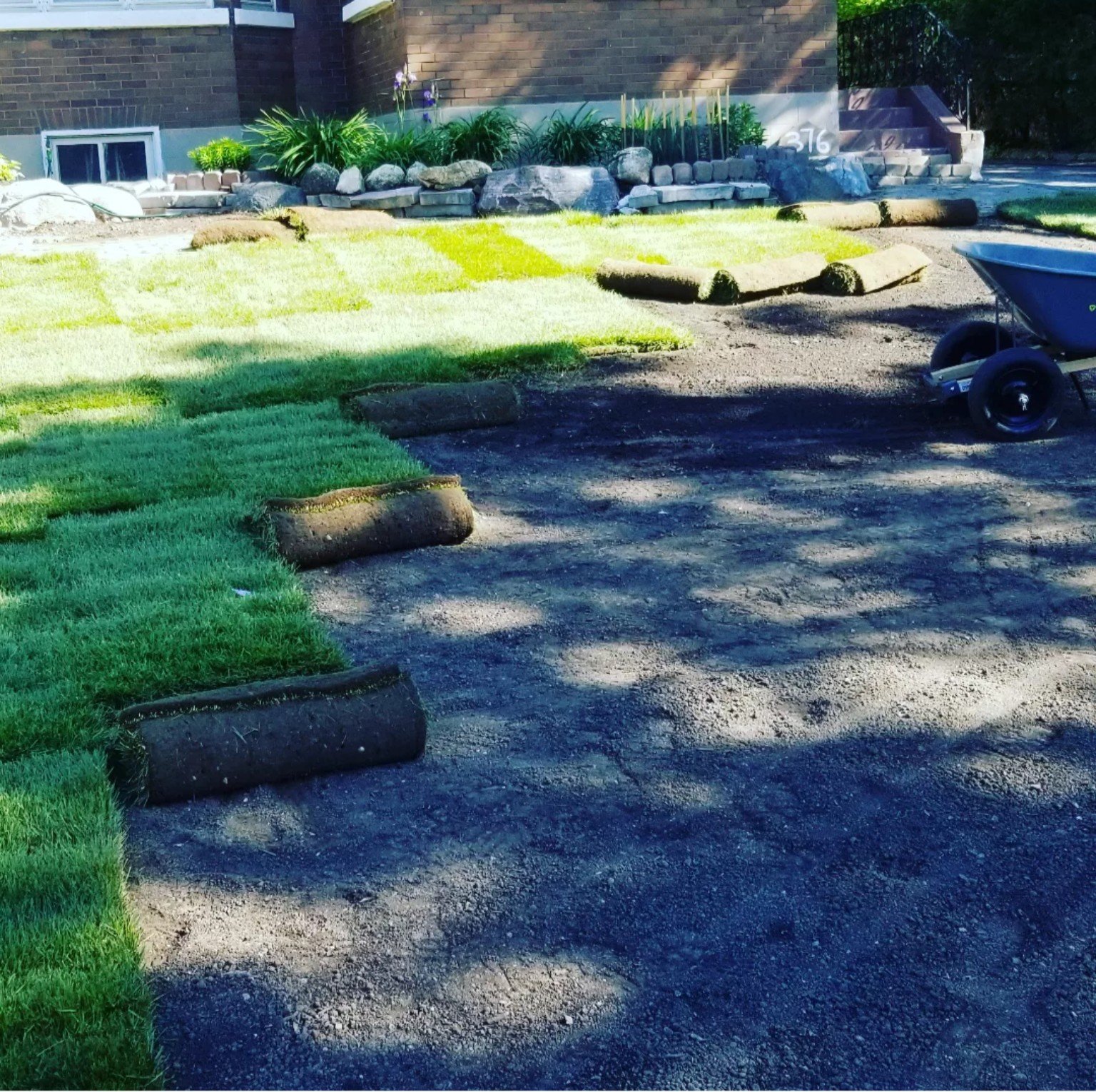 Backyard garden with newly laid sod, landscaping stones, and a wheelbarrow on dirt path. House with brick exterior and stairs in background.