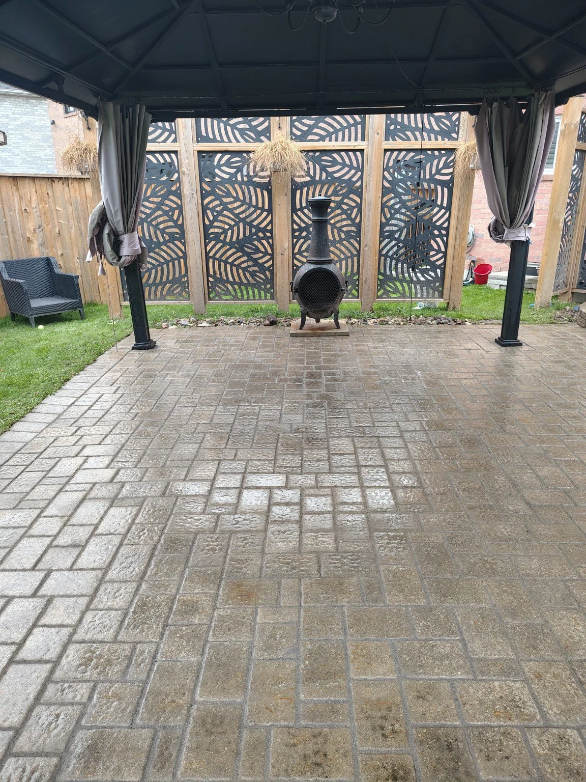 A covered outdoor patio with a brick floor and a decorative metal privacy screen at the back. There is a black outdoor heater or stove centered against the screen. To the left is a black outdoor chair on a small grassy area, and to the right is a red