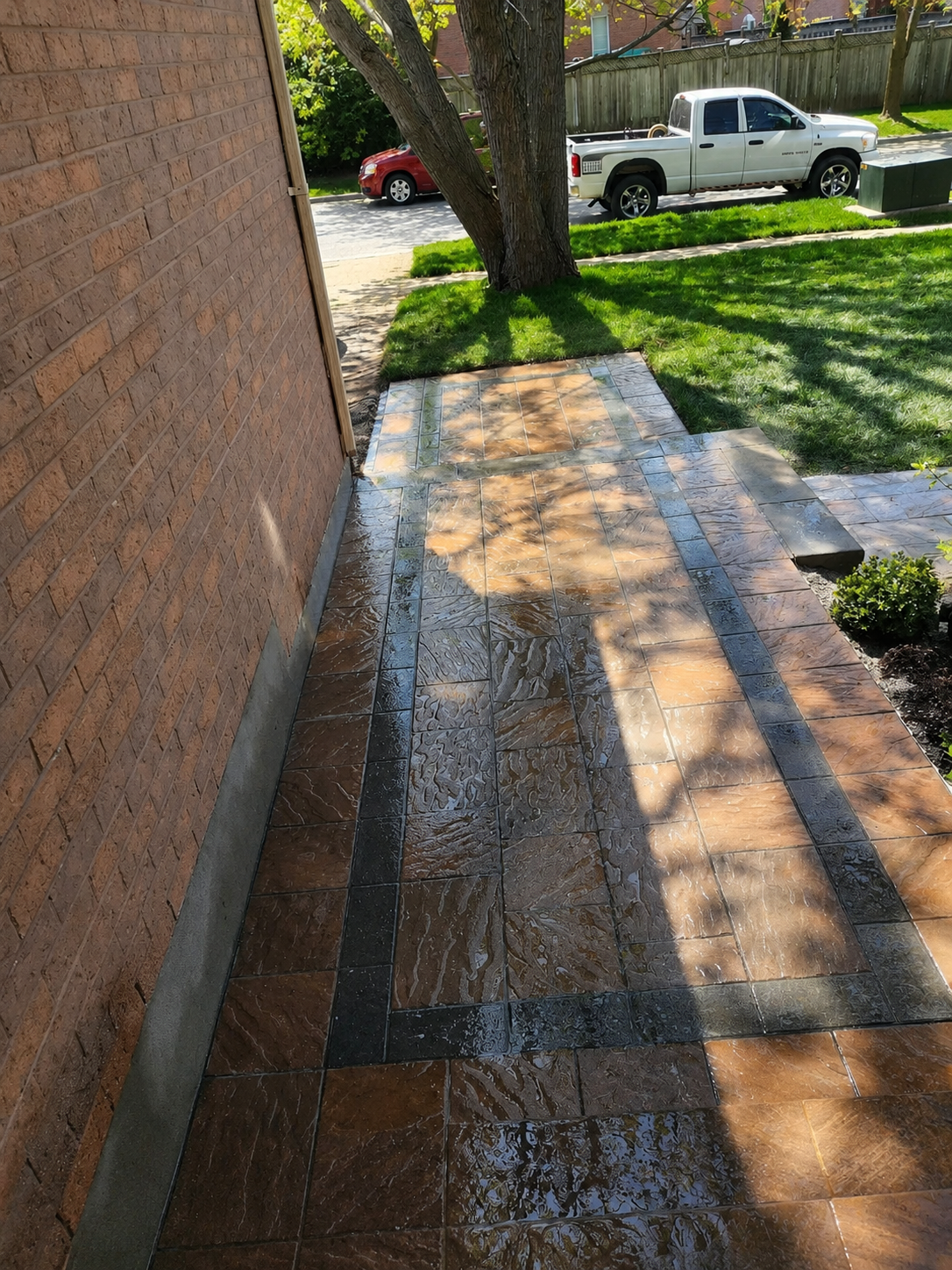 Wet brown and black tiled walkway beside a brick house with a tree, green grass, and parked vehicles in the background.
