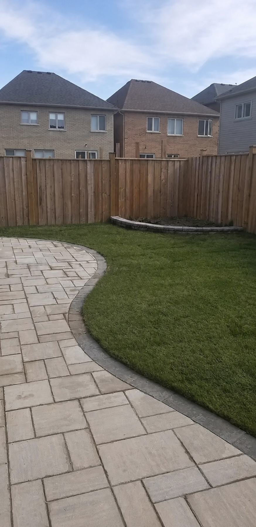 A backyard with a paved stone patio, a curved grass lawn, a fenced yard with a small garden bed, and neighboring houses in the background under a partly cloudy sky.