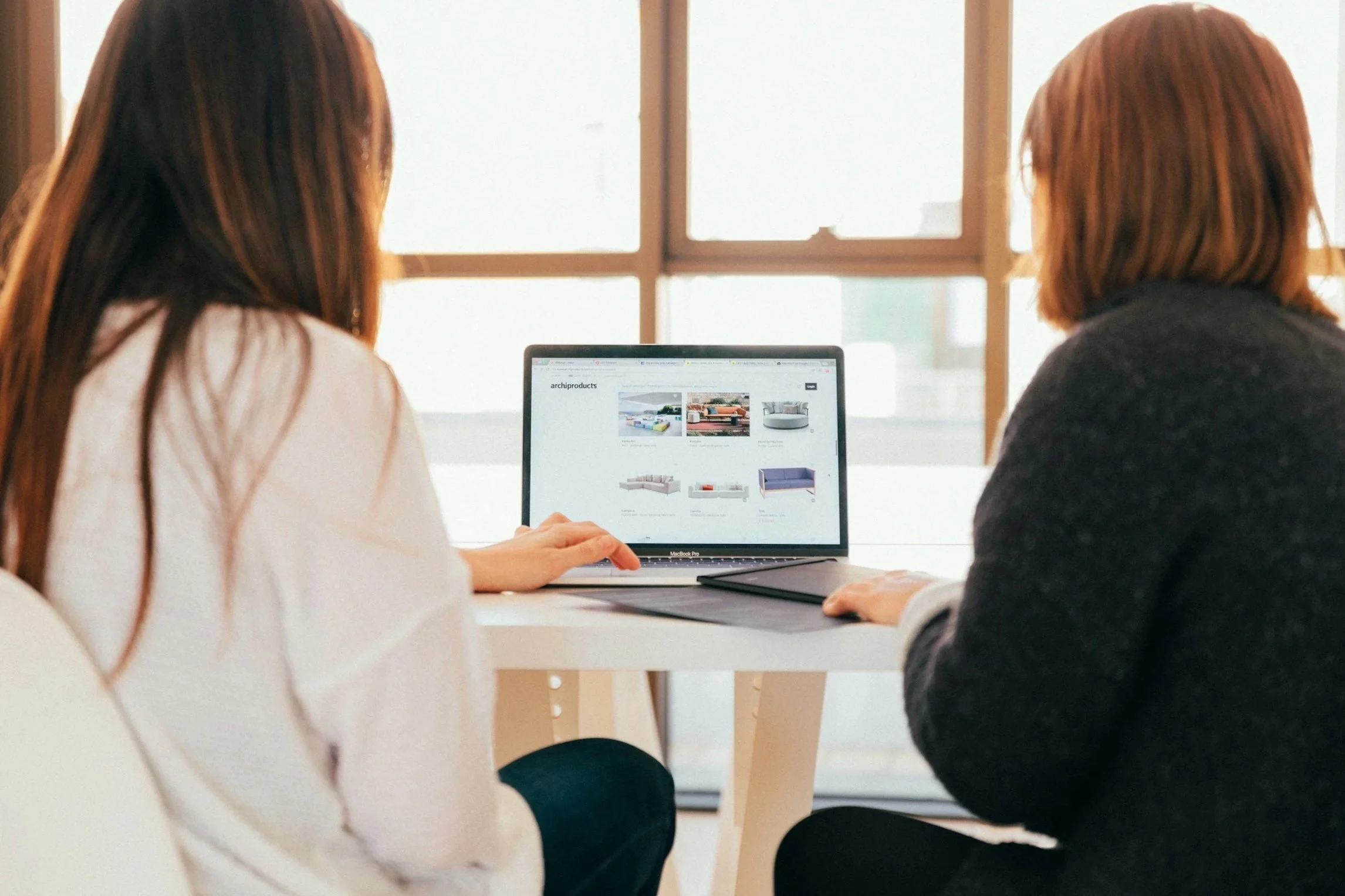 Dos mujeres sentadas en una mesa frente a una computadora portátil, explorando un sitio web de muebles para el hogar en un espacio con ventanas grandes y luz natural.