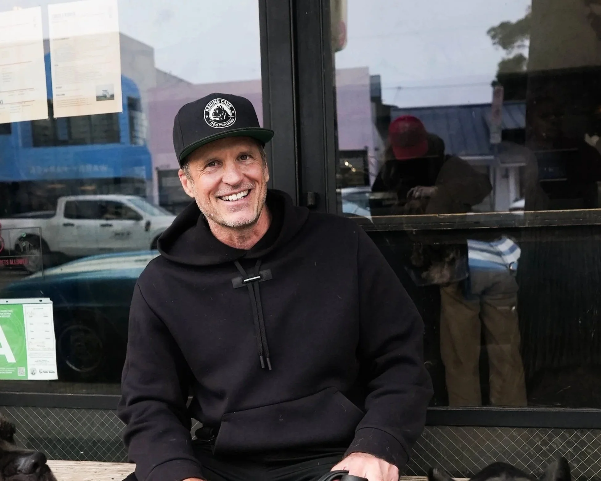 A smiling man in a black hoodie and a black cap with a dog training logo is sitting outside a storefront with large glass windows, behind him various cars are parked and a person wearing a red cap can be seen in the reflection.