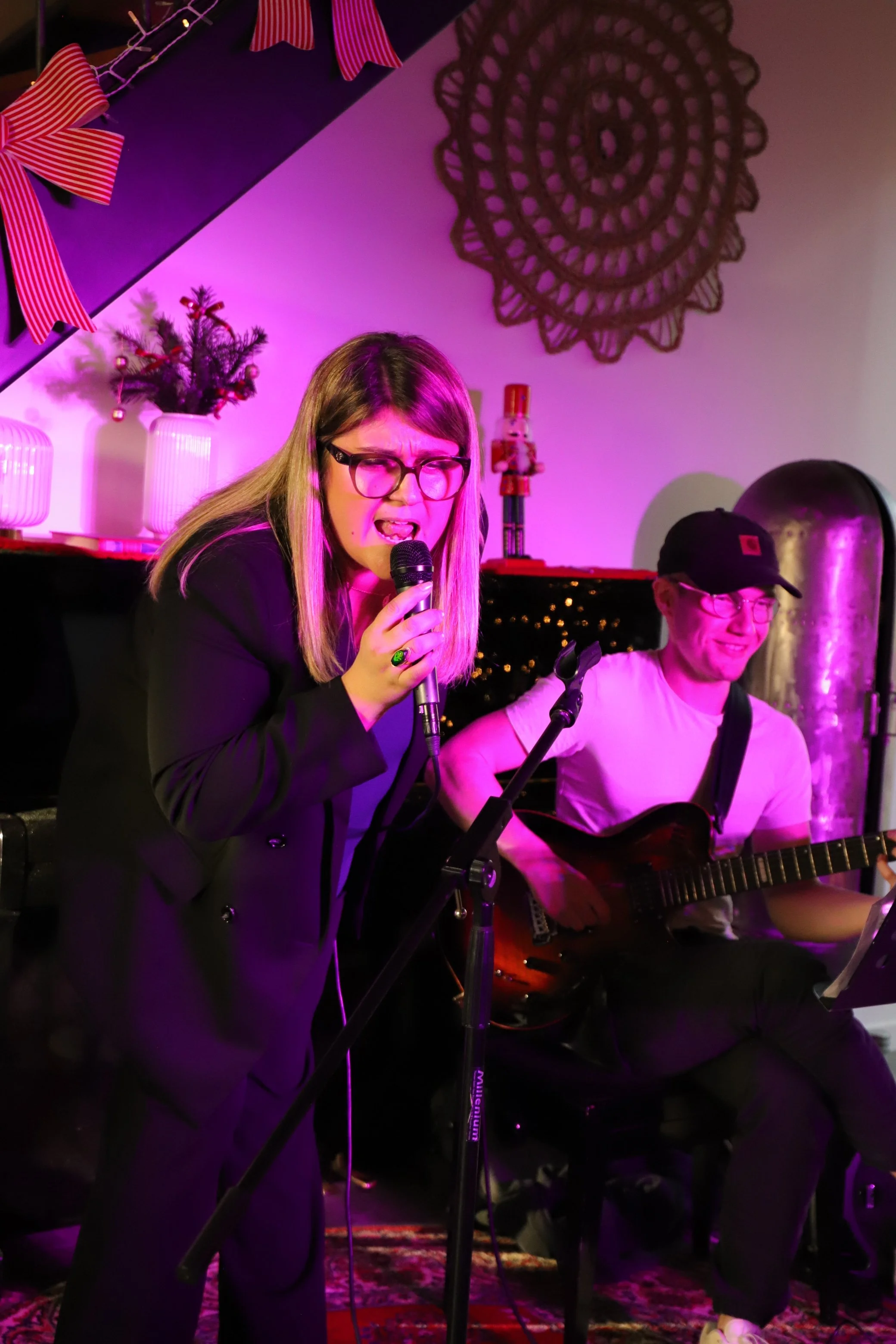 Deux personnes jouent de la musique dans une pièce décorée pour Noël, avec un sapin miniature, des rubans rouges et blancs, et une décoration murale en crochet noire sous une lumière violette.