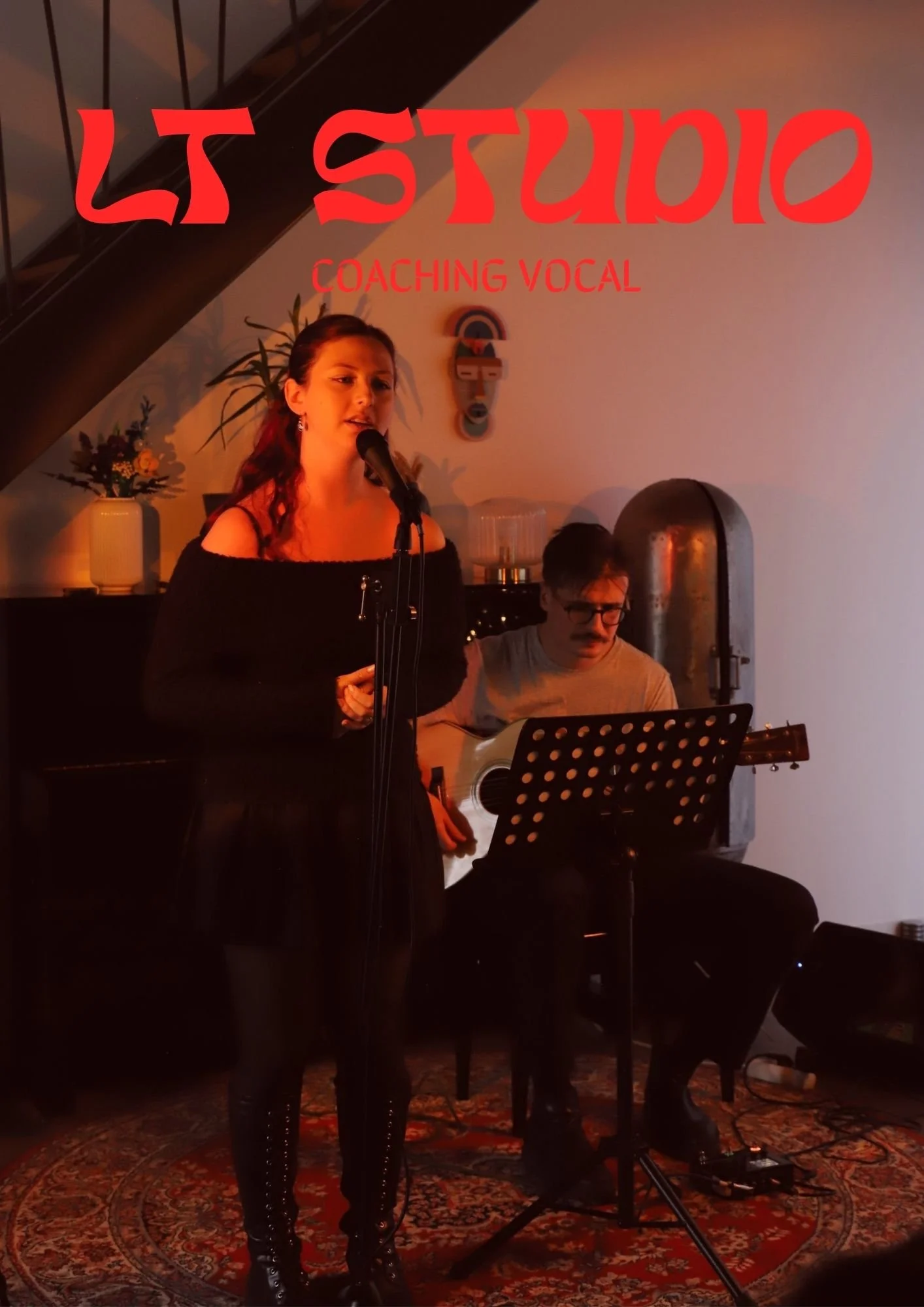 Une femme chante devant un microphone, accompagnée d'un guitariste. La scène est interior avec une décoration chaleureuse et un tapis devant eux.