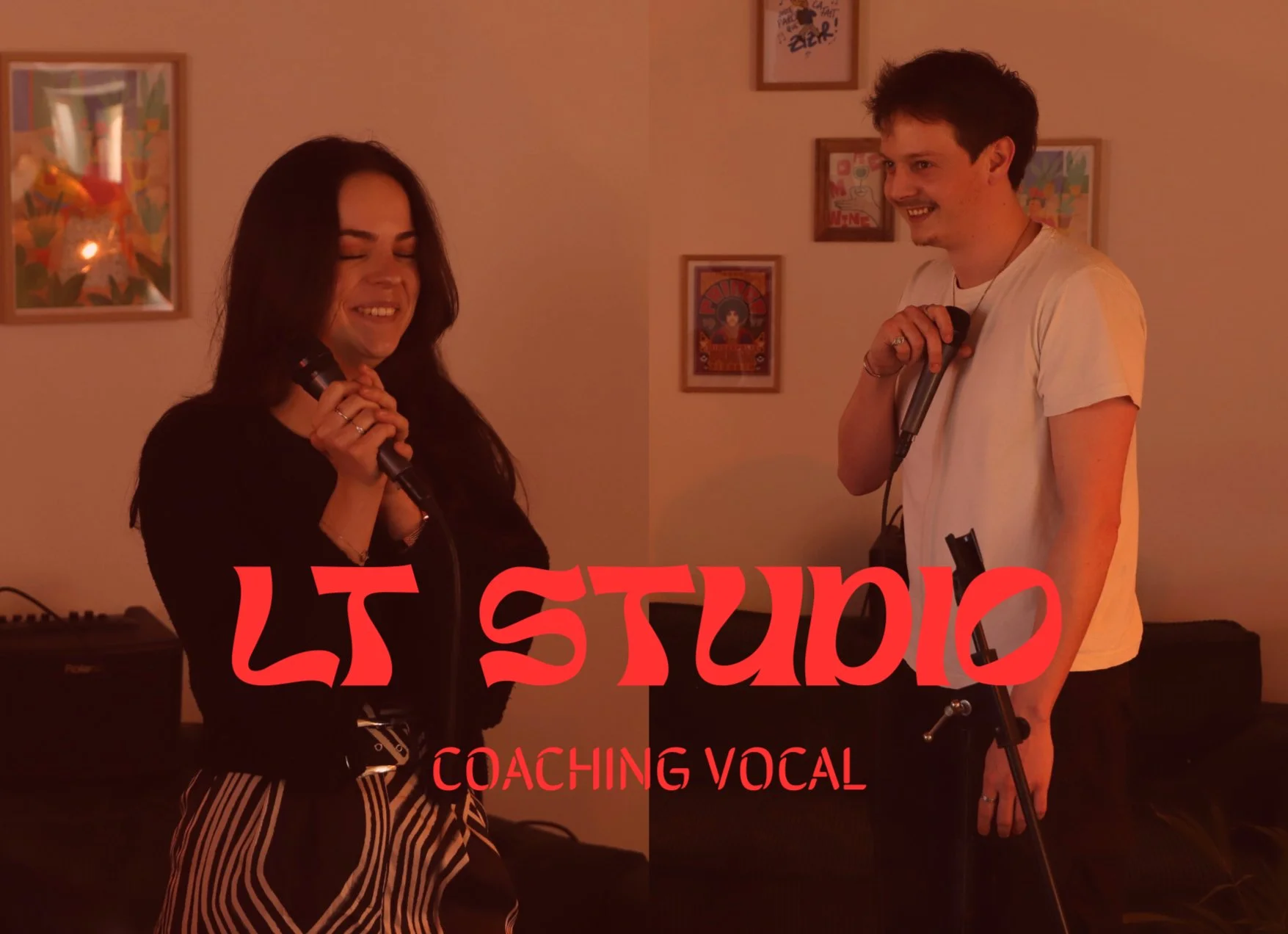Deux personnes, un homme et une femme, chantent ou parlent dans une pièce, chacun tenant un microphone. L'arrière-plan comprend des œuvres d'art encadrées sur le mur et un clavier électronique. Le texte overlay en rouge dit "LSTUDIO" et "COACHING VOCAL".
