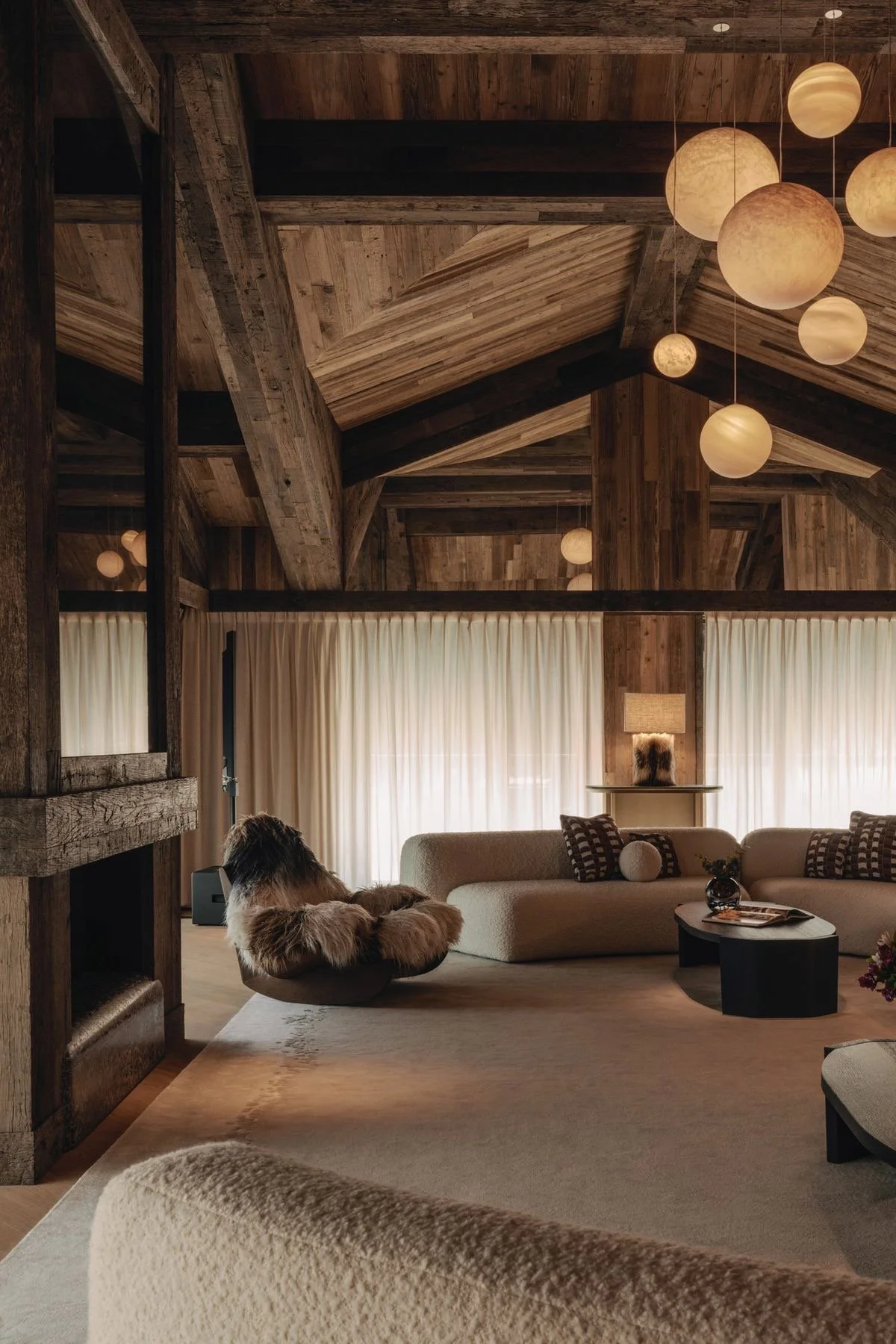Interior of a cozy living room with wooden ceiling beams and hanging spherical pendant lights, beige curtains, a light-colored sofa with patterned pillows, a side table with a lamp and fur decoration, and a few decorative items on a coffee table.