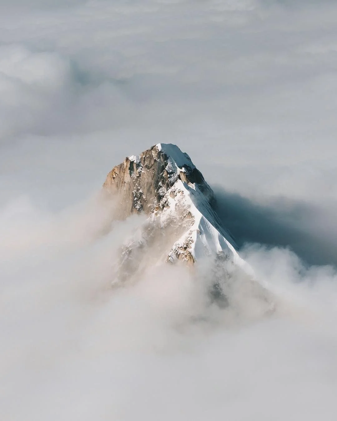 Snow-covered mountain peak rising above clouds with blue sky and wispy clouds above.