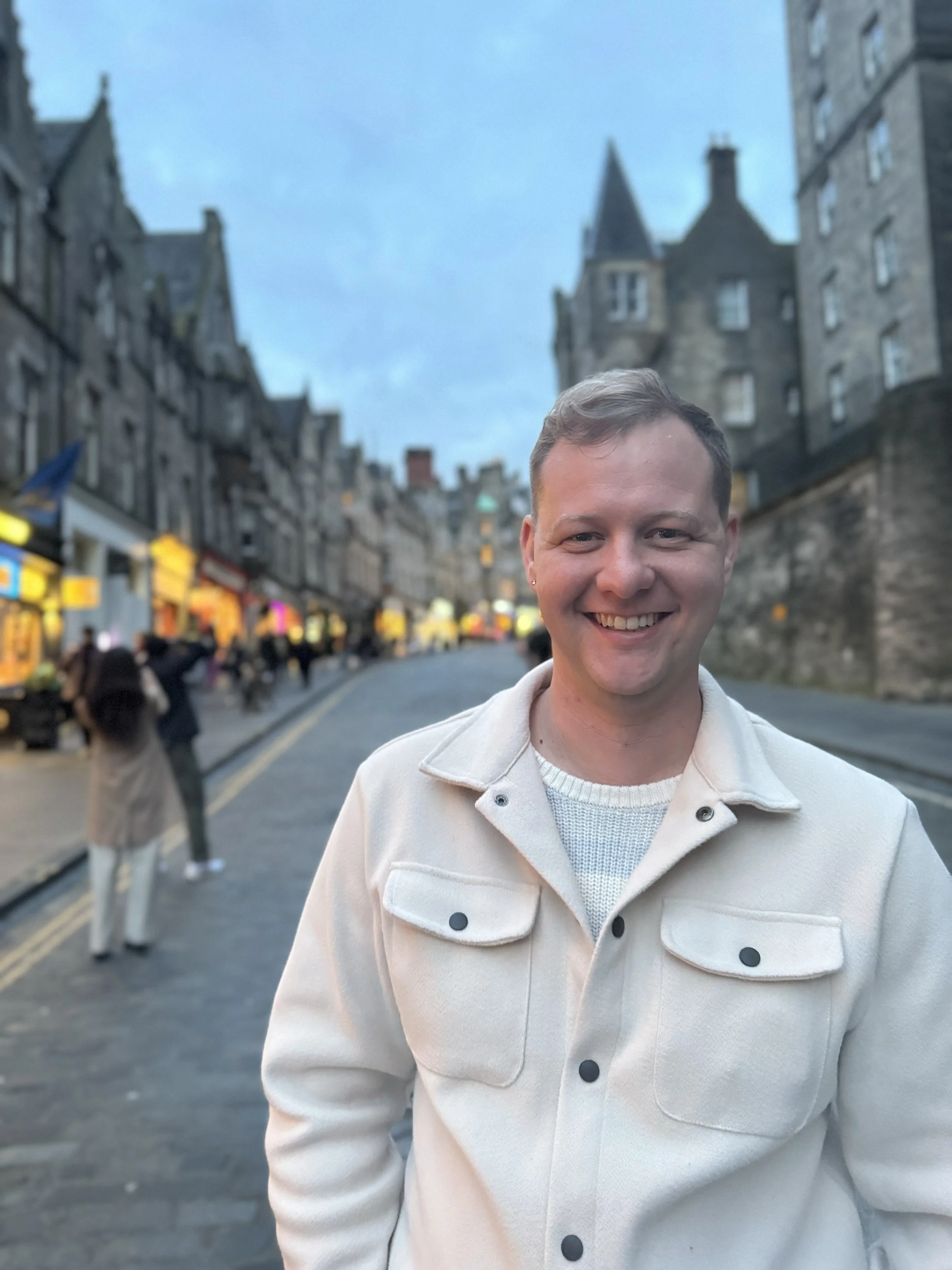 Smiling person in a winter coat standing on a busy street with historic buildings and people in the background.