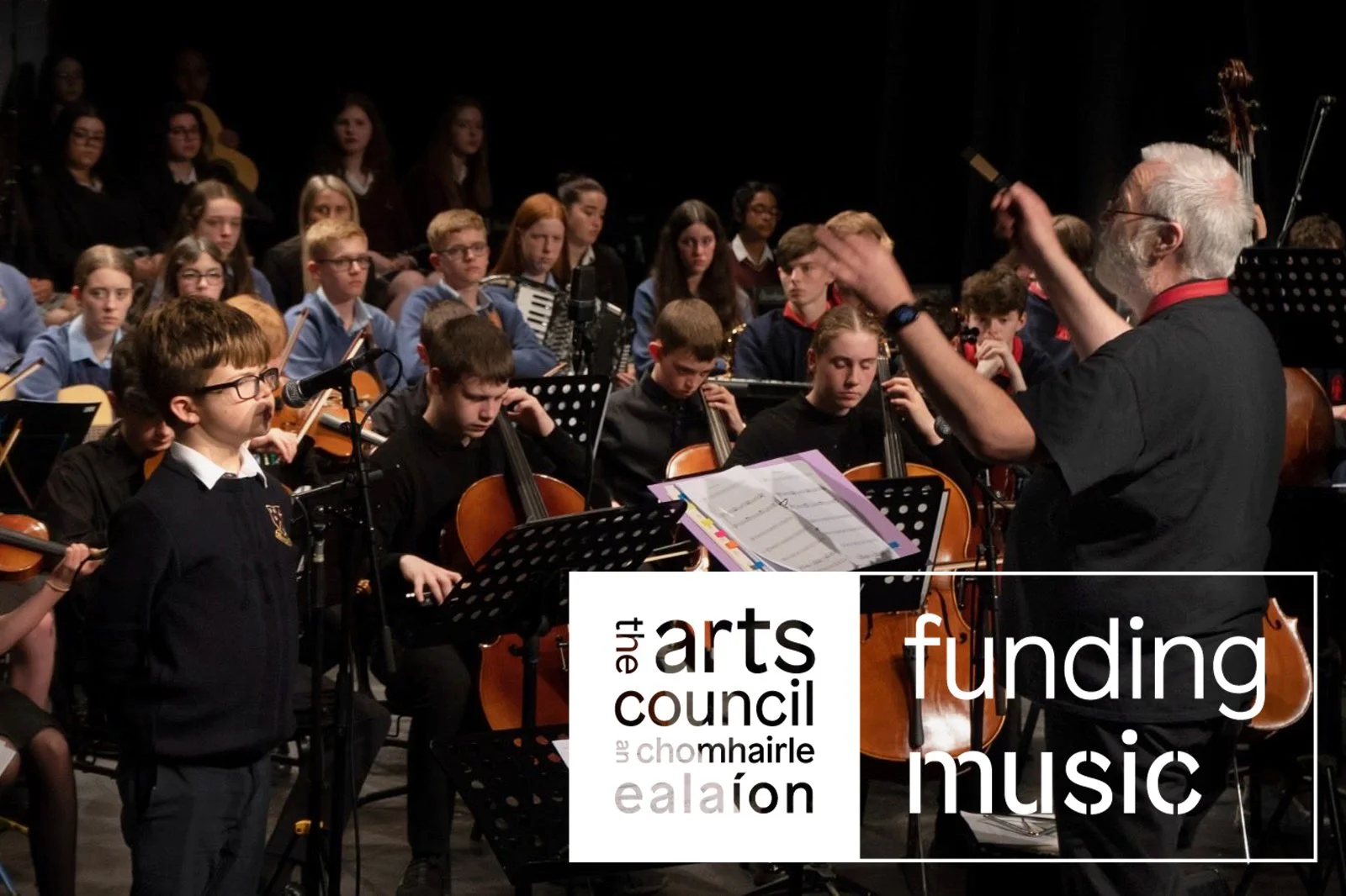 Quartet member Kenneth Edge conducts an orchestra comprised of young musicians. On the left of the image we see a young person with glasses singing as a soloist.