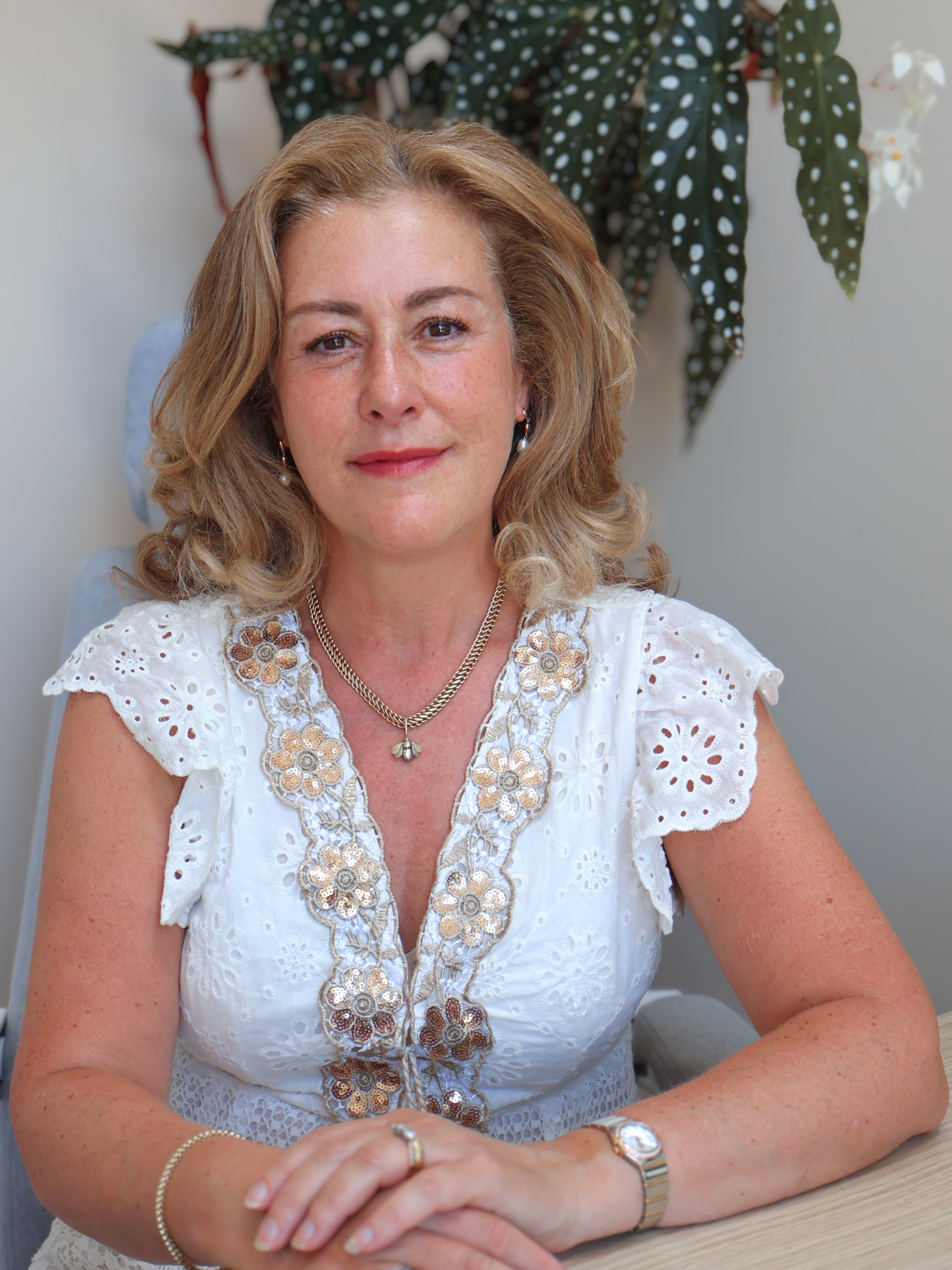 A woman with blonde, styled hair, wearing a white eyelet lace top decorated with flower embroidery, gold jewelry, including earrings, a necklace, and a bracelet, is sitting at a desk. Behind her is a plant with green leaves with white spots and white flowers.