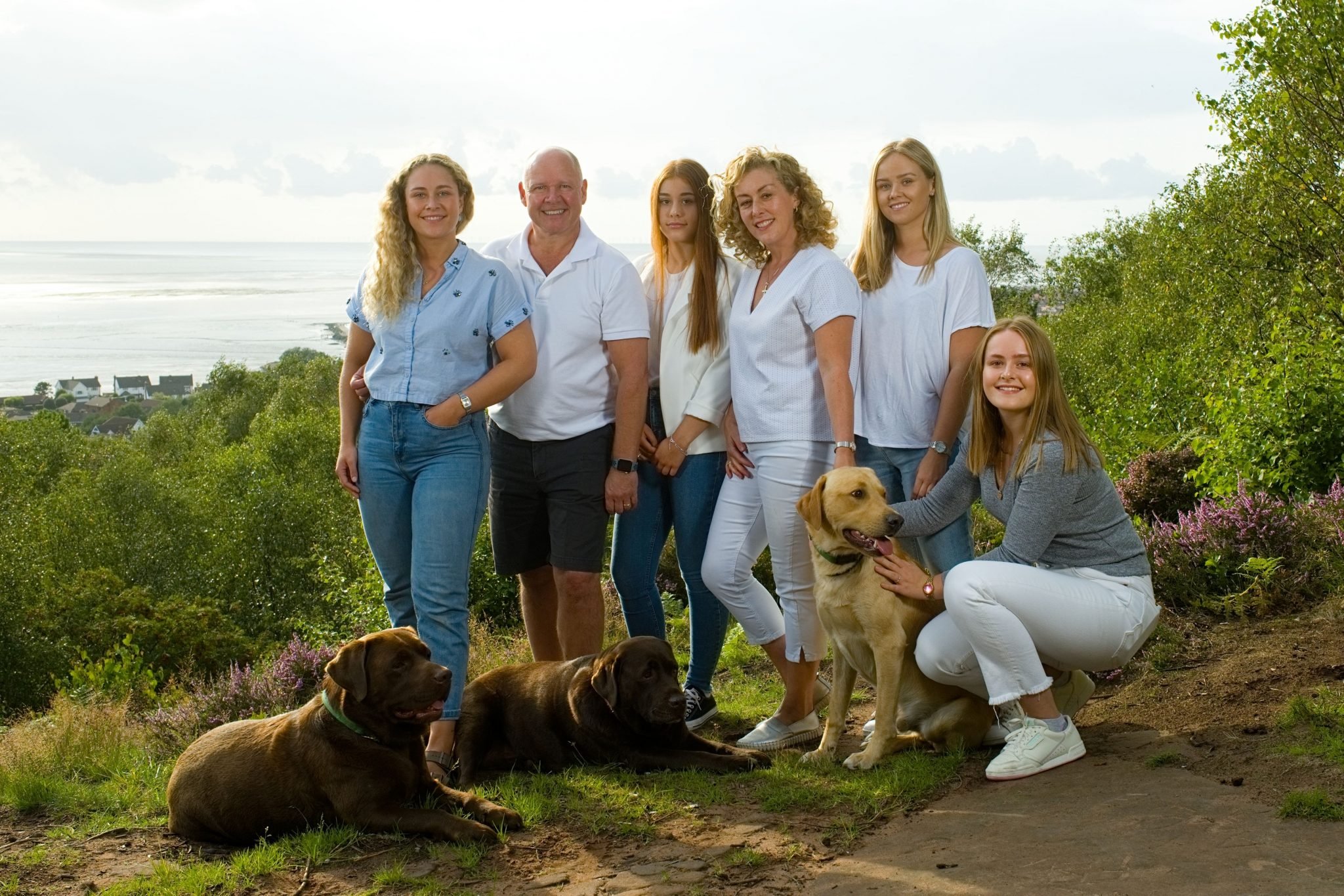 A group of seven people and three dogs outdoors near the coast, smiling and posing for a photo, with greenery and trees around.