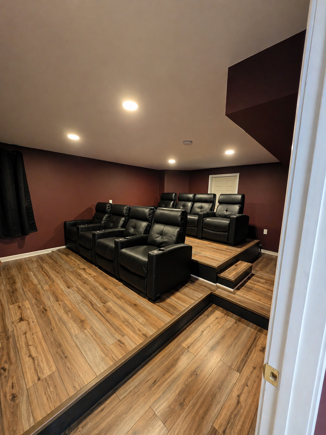 Finished basement theater room with tiered seating, black leather recliners, wood-look flooring, recessed lighting, and dark cinematic walls.