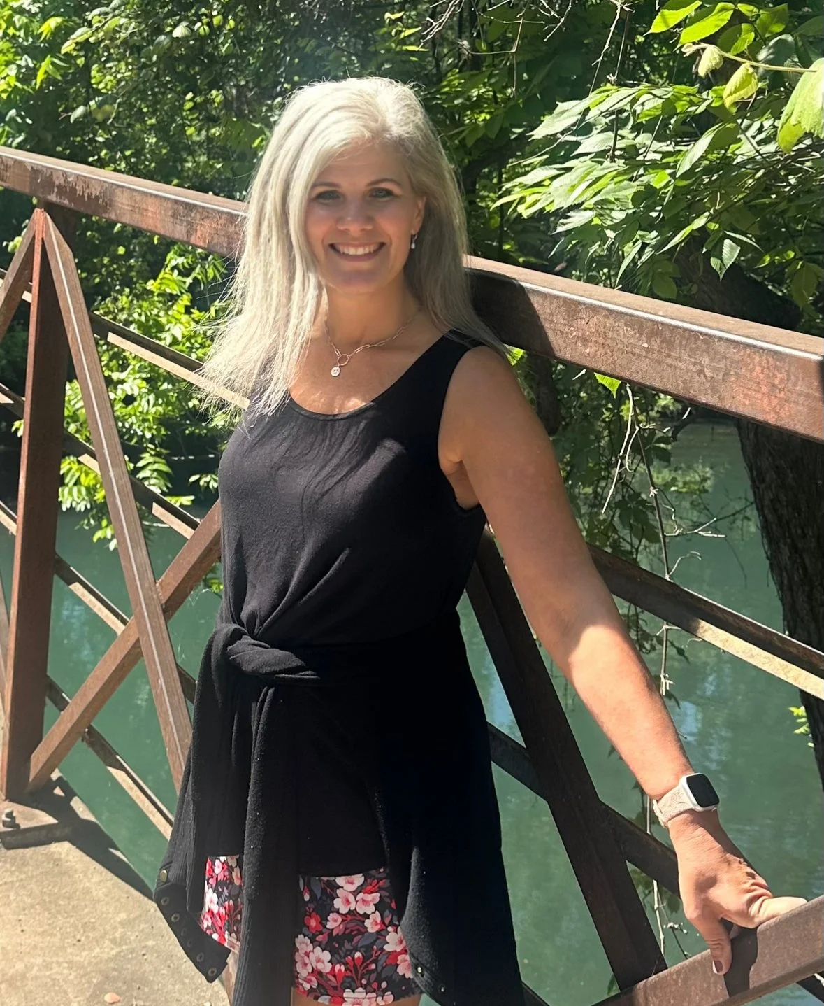 A woman with blonde hair smiling and standing outdoors near a body of water and green trees, wearing a black sleeveless dress with a tied waist, floral patterned shorts, a necklace, and a smartwatch, on a metal bridge.