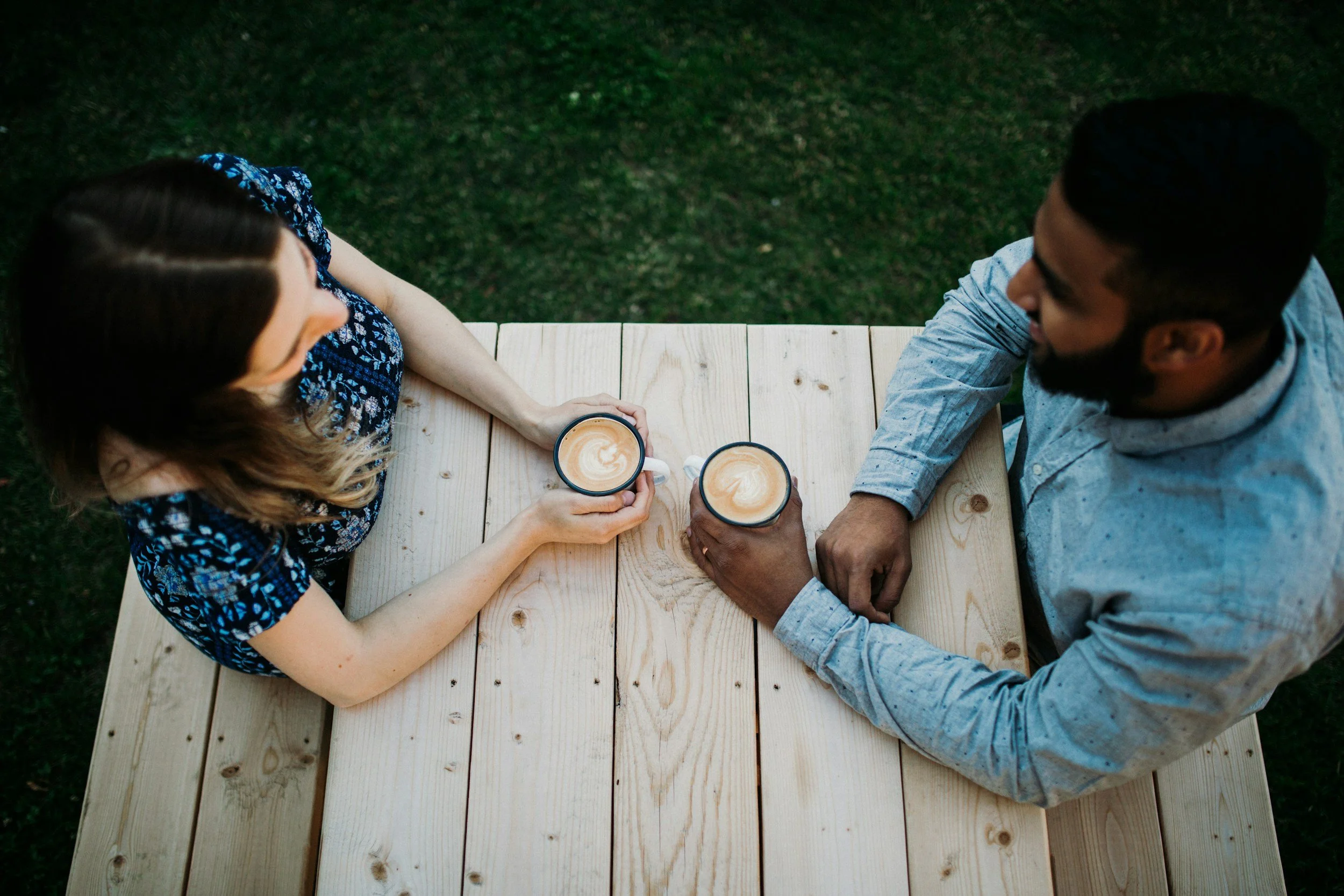 Two people sitting at a light-colored wooden table outdoors, holding cups of coffee or latte, viewed from above.