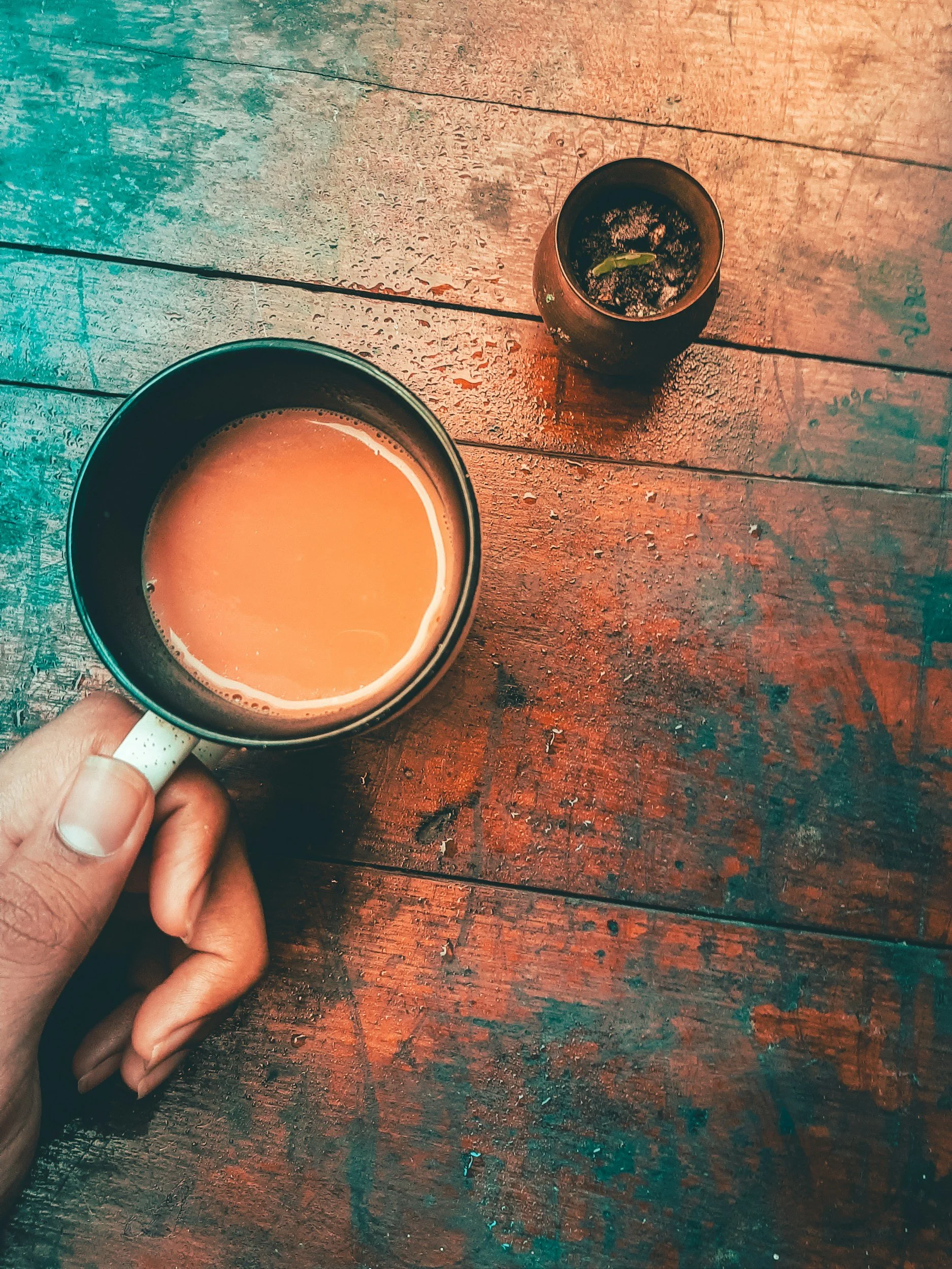 A person's hand holding a mug filled with coffee or tea, on a wooden table. Next to the mug, there is a small round container with some green leaves or herbs inside.
