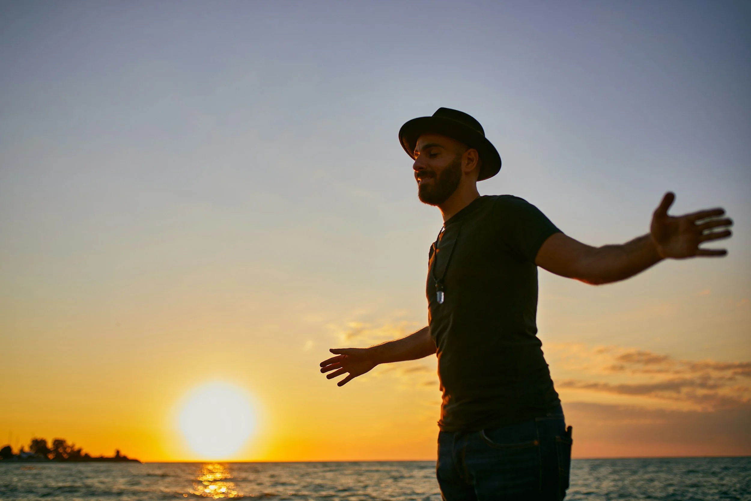 A man with a beard wearing a black hat and black shirt is standing by the water at sunset, with his arms outstretched.