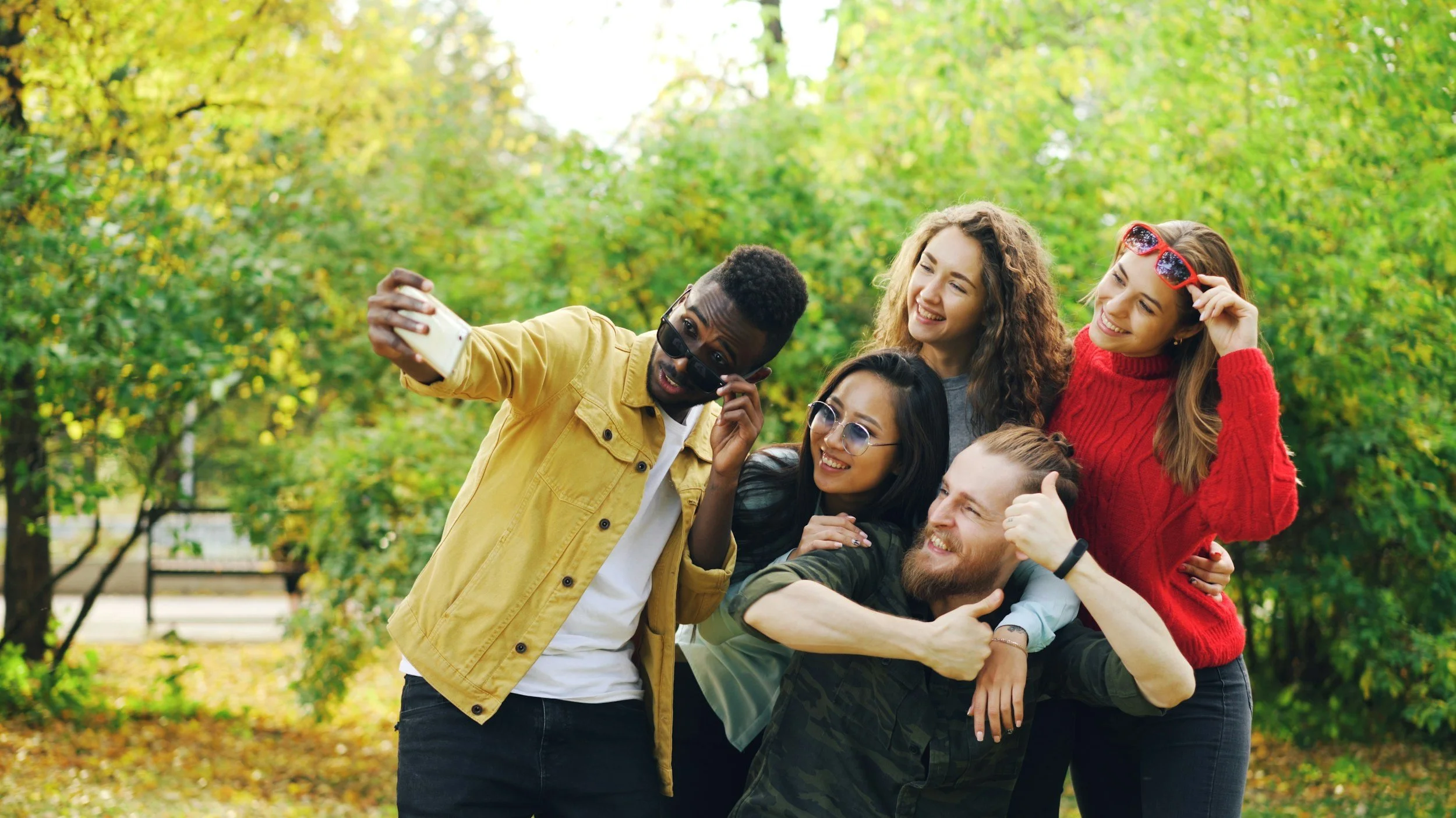 Group of five friends taking a selfie outdoors in a park with green trees. They are smiling and appear happy.