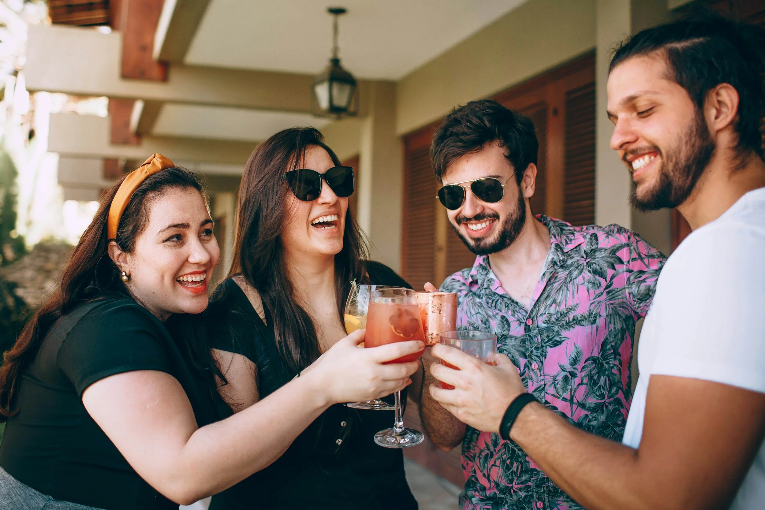 A group of four friends at an outdoor gathering, holding colorful drinks and smiling, with a woman wearing sunglasses and a man in a pink Hawaiian shirt in the center.