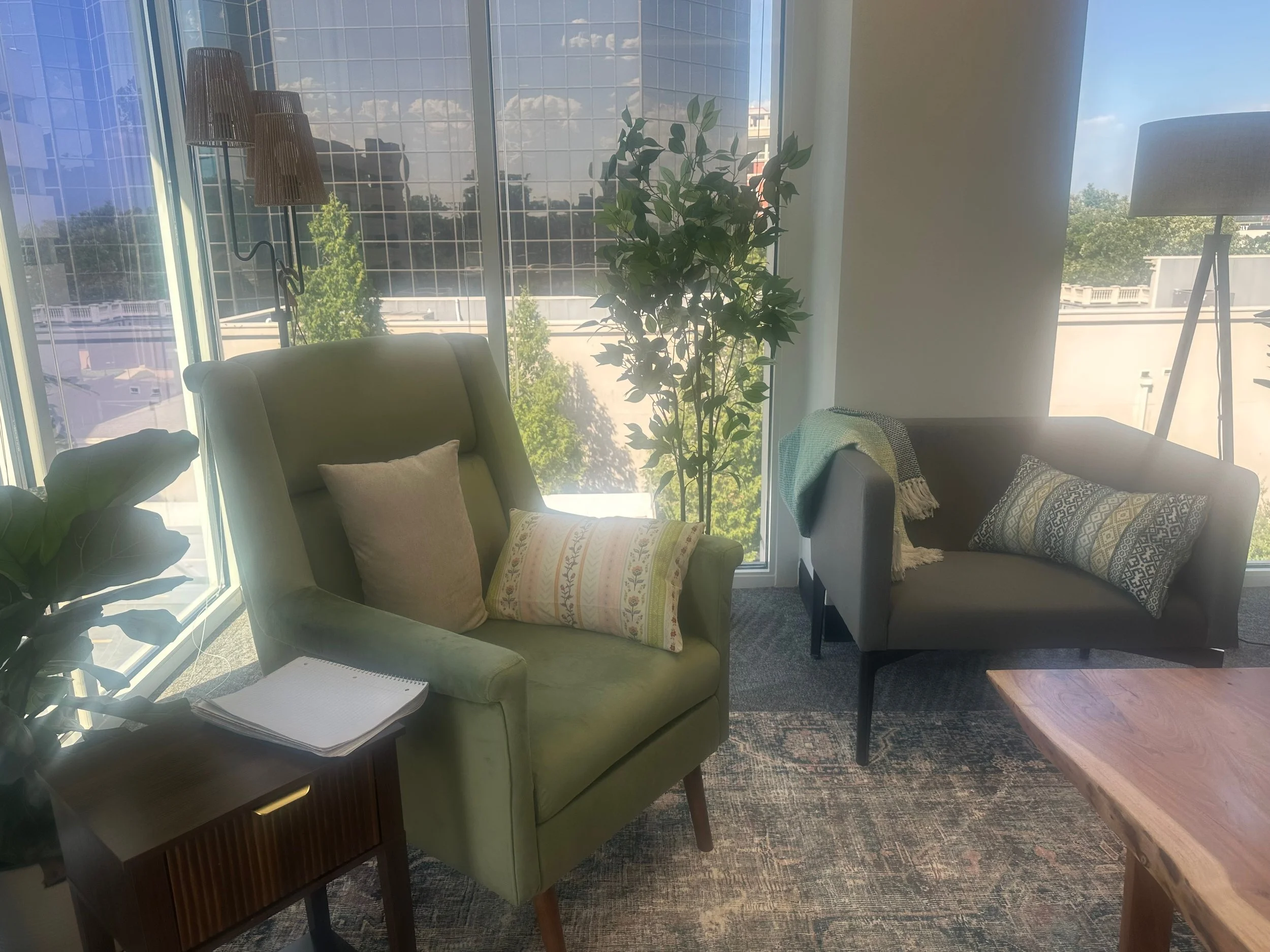 Interior of a modern room with large floor-to-ceiling windows, a beige armchair with decorative pillows, a small side table with a notepad, a tall plant near the window, a wooden coffee table, a tripod floor lamp and a neighboring building visible through the windows.