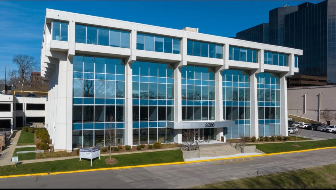 Modern office building with glass windows and geometric white structural elements at 3200, with a parking lot and landscaped area in front.