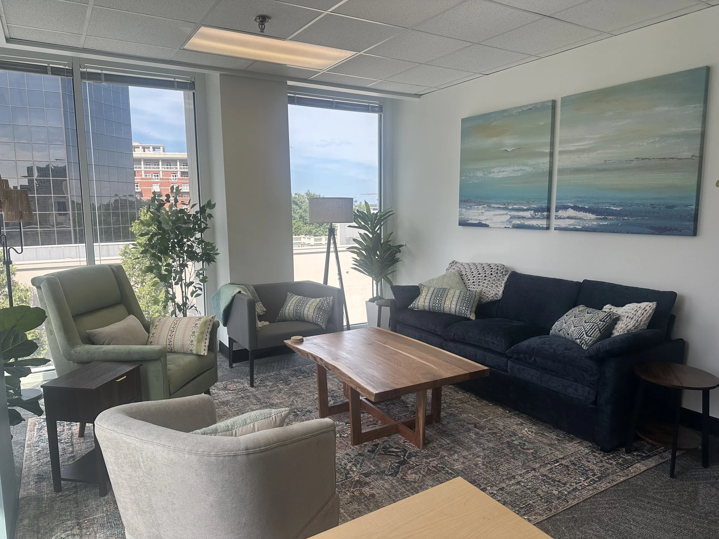 A modern office lounge area with large windows showcasing a city view. The space is decorated with multiple green and beige armchairs, a black sofa with patterned pillows, small tables, a wooden coffee table, and potted plants. There are two large paintings of ocean scenes on the white wall.