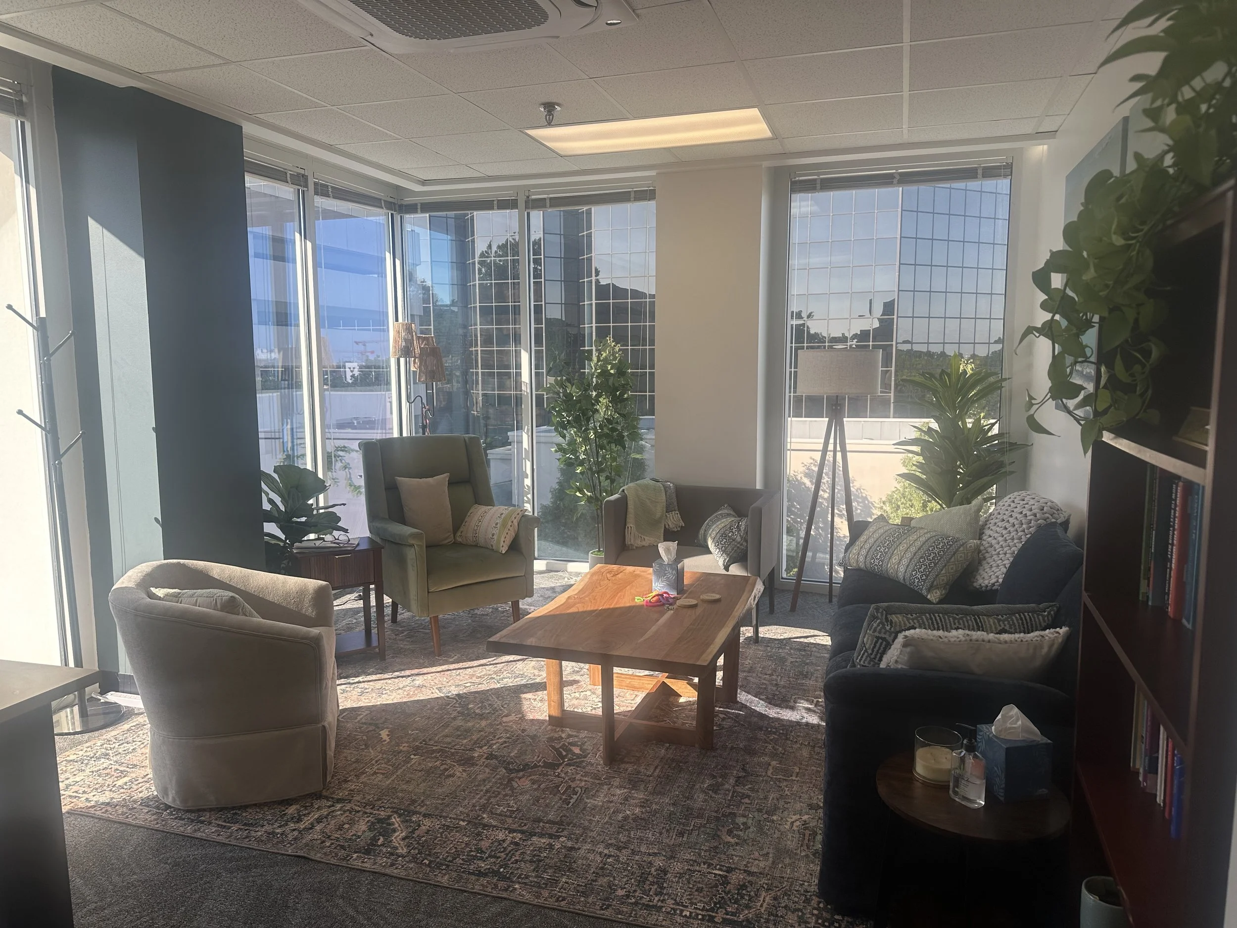 A modern office lounge with large windows allowing natural light. There are armchairs, a small coffee table, plants, and a bookshelf, creating a cozy and inviting space.