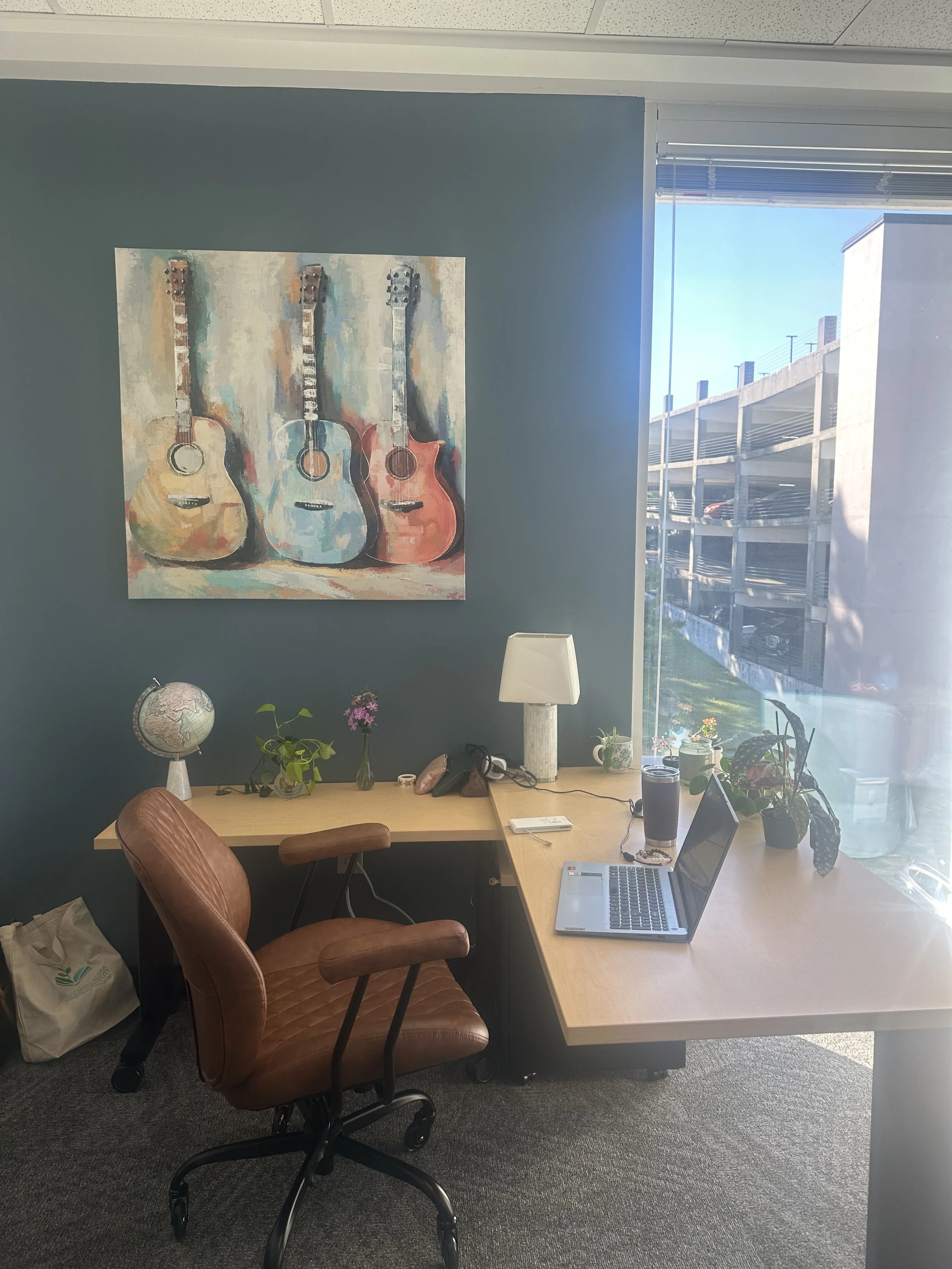 An office workspace with a wooden desk, a brown leather office chair, a laptop, a lamp, various plants, and a painting of three guitars hanging on a teal-colored wall. There is a window with a view of an outdoor parking structure.