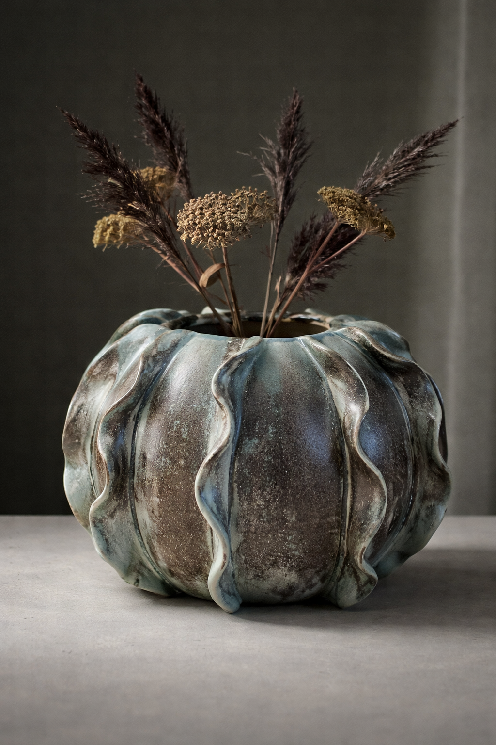 En keramikvas med vågformade kanter, fylld med torkade blommor och gräs. Vasen har en blågrå yta med en glansig finish, placerad på en ljus yta mot en mörk bakgrund.