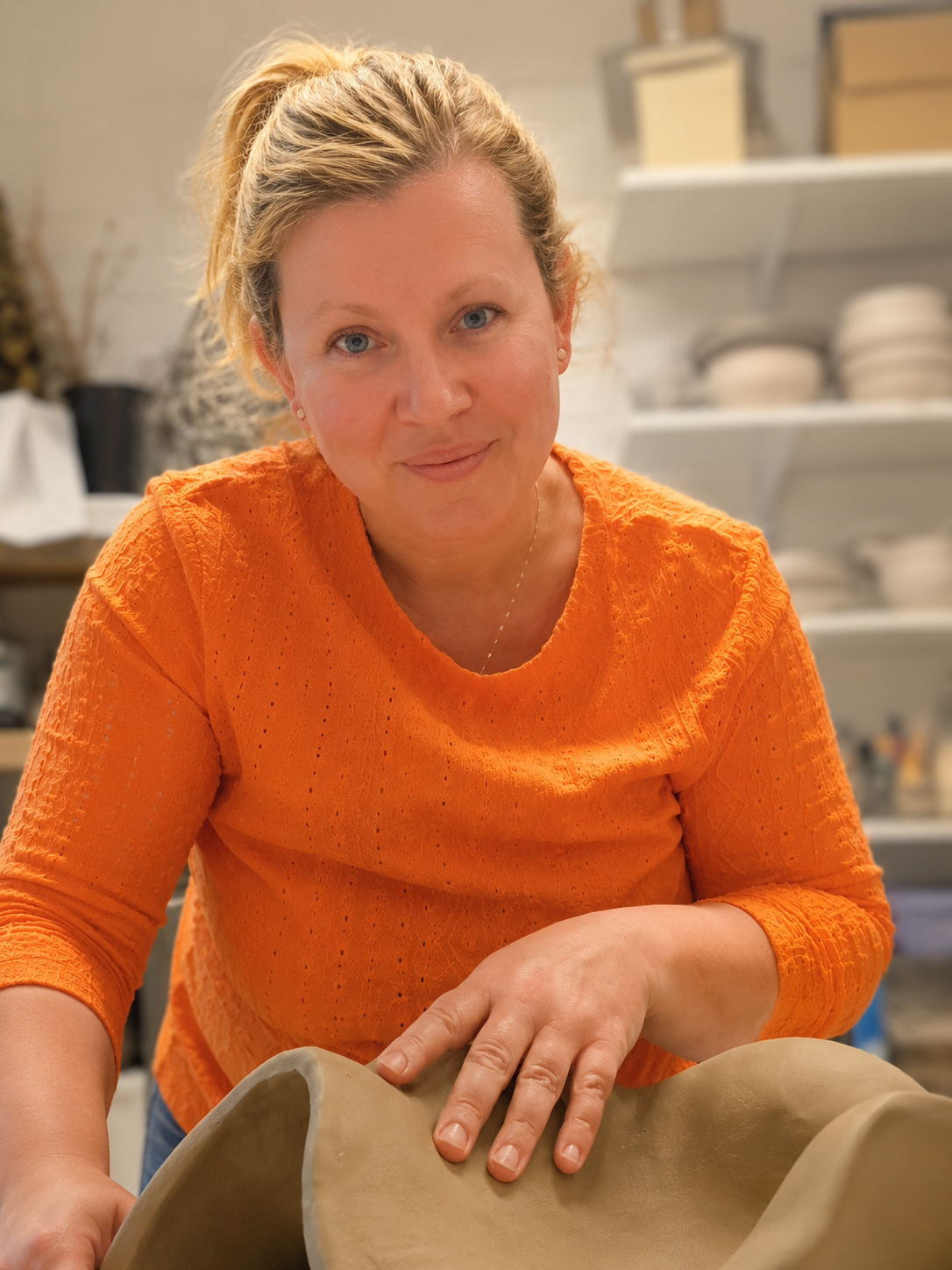 Kvinna som arbetar med lera i ett verkstadsutrymme, bär en orange tröja och formar leran.