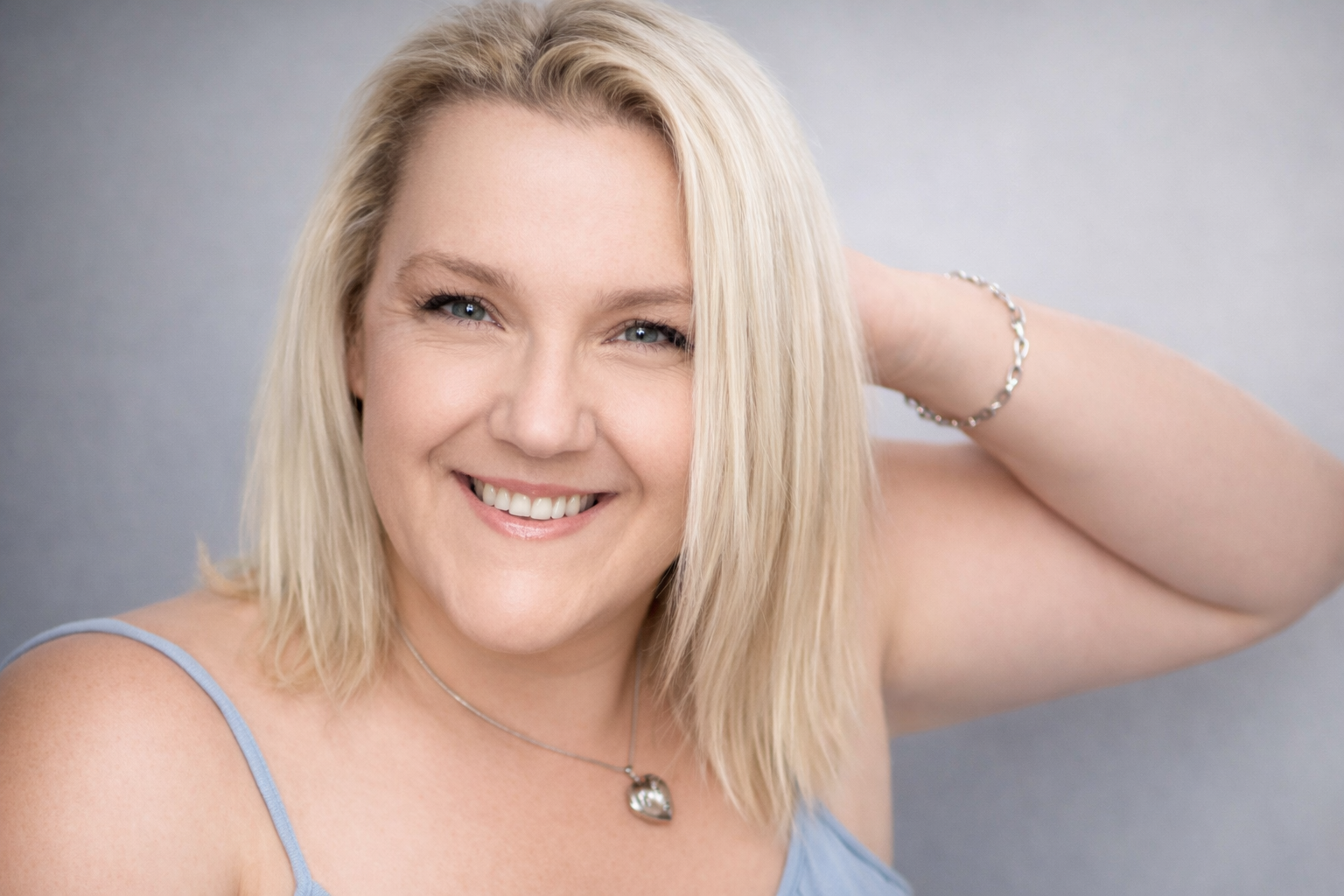 A smiling blonde woman with shoulder-length hair, wearing a blue tank top, a silver bracelet, a necklace, and looking at the camera against a neutral background.