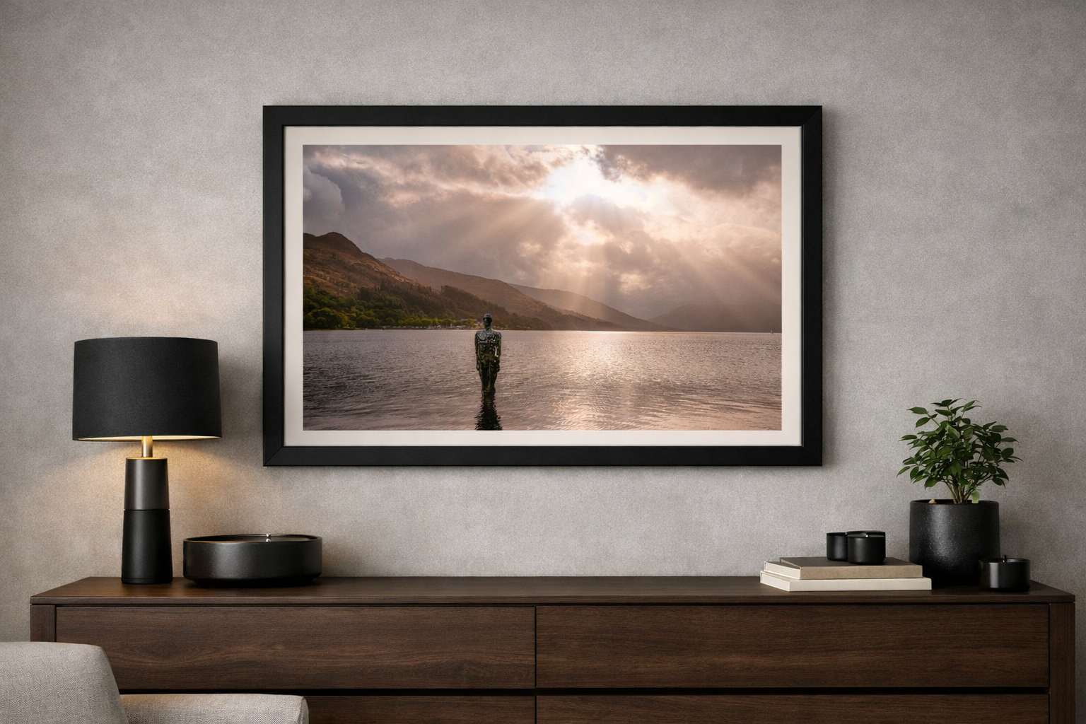 Living room with a framed landscape photo of a person standing in water with mountains and cloudy sky, a black table lamp, black decor items, a small plant, and books on a wooden sideboard.